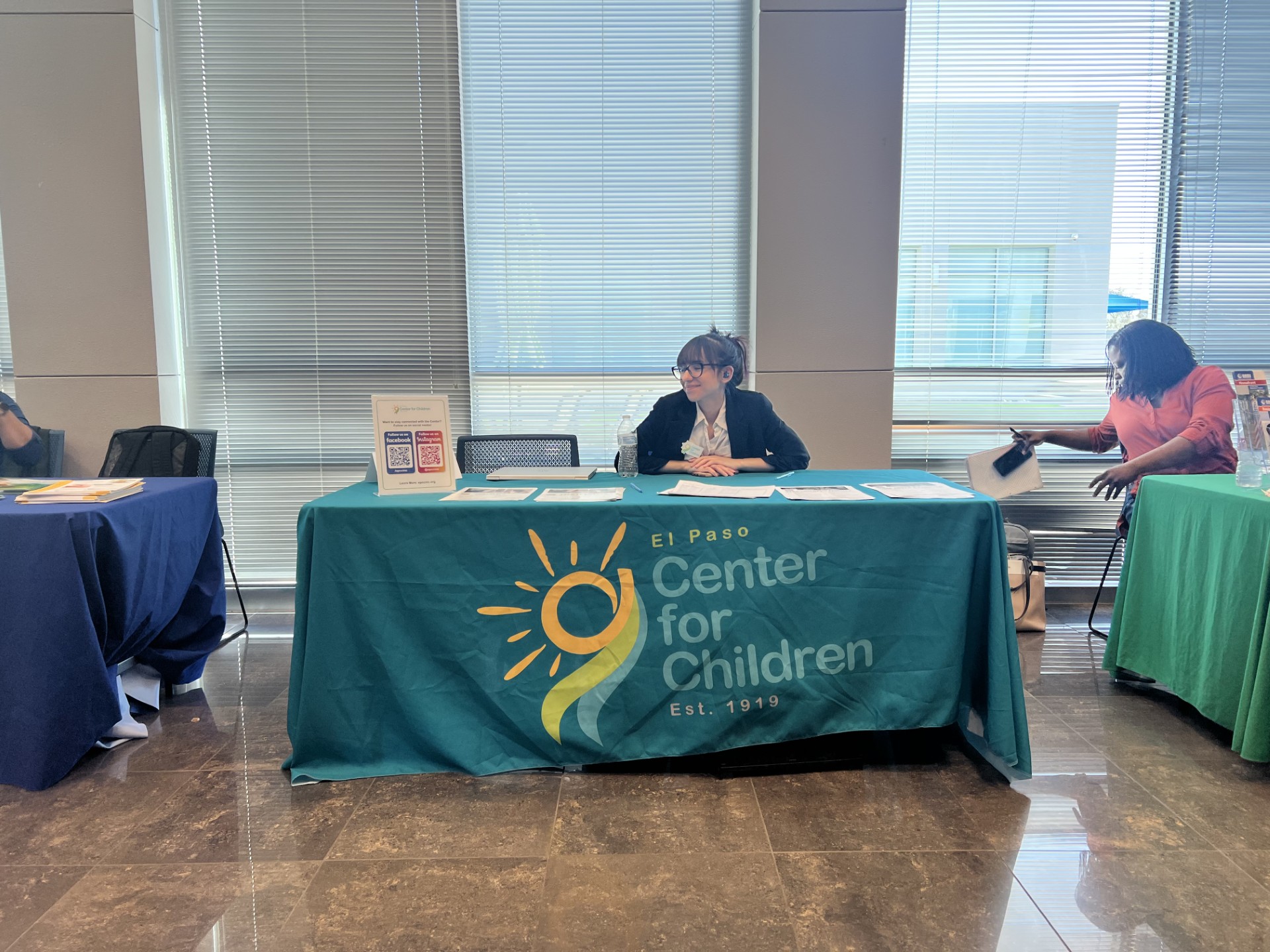 A teal tablecloth with "El Paso Center for Children" printed on it is in the foreground, while a woman in an orange shirt is visible in the background.