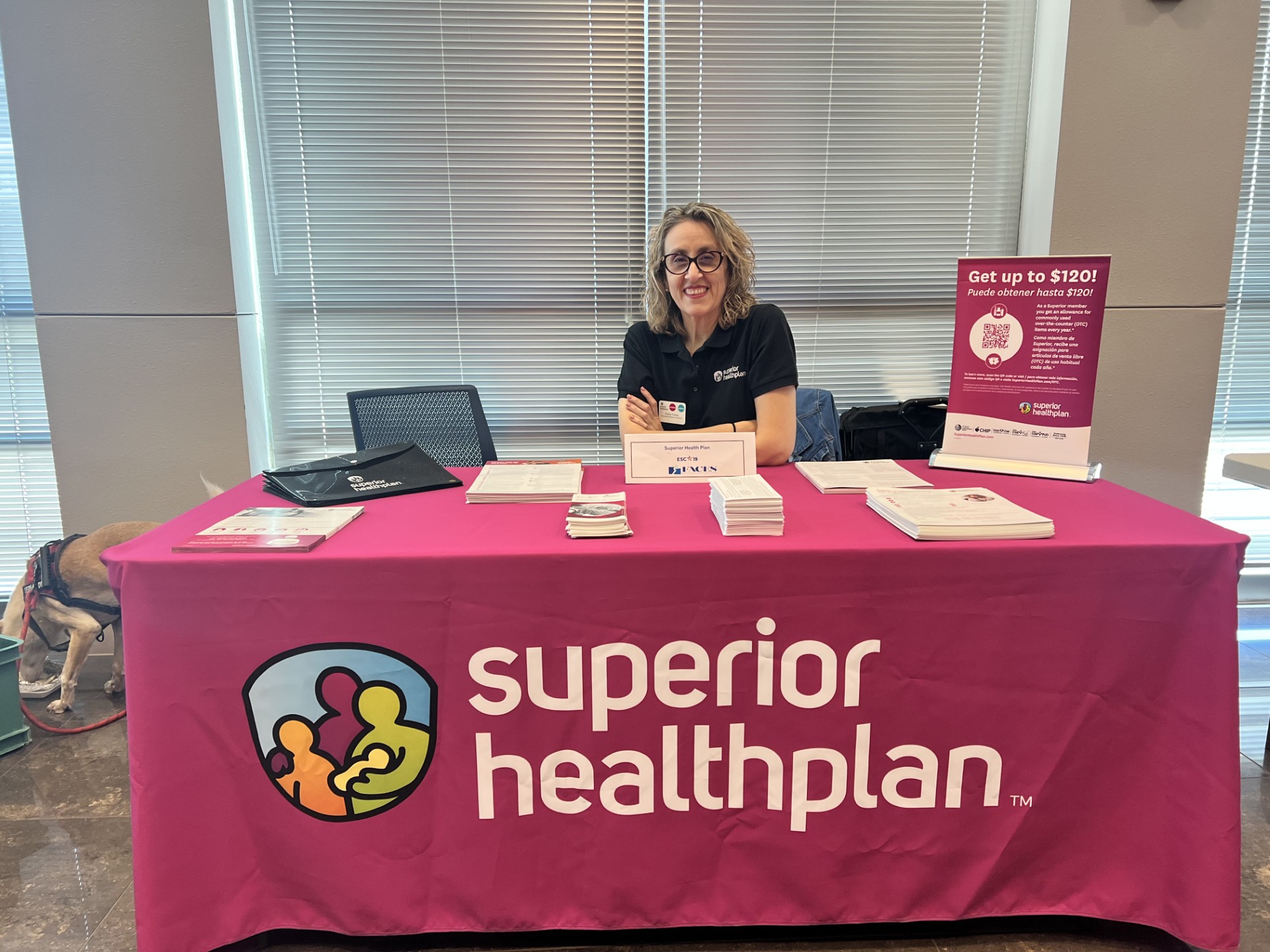 A woman smiles behind a table with "superior healthplan" branding, while blinds and a window form the background.