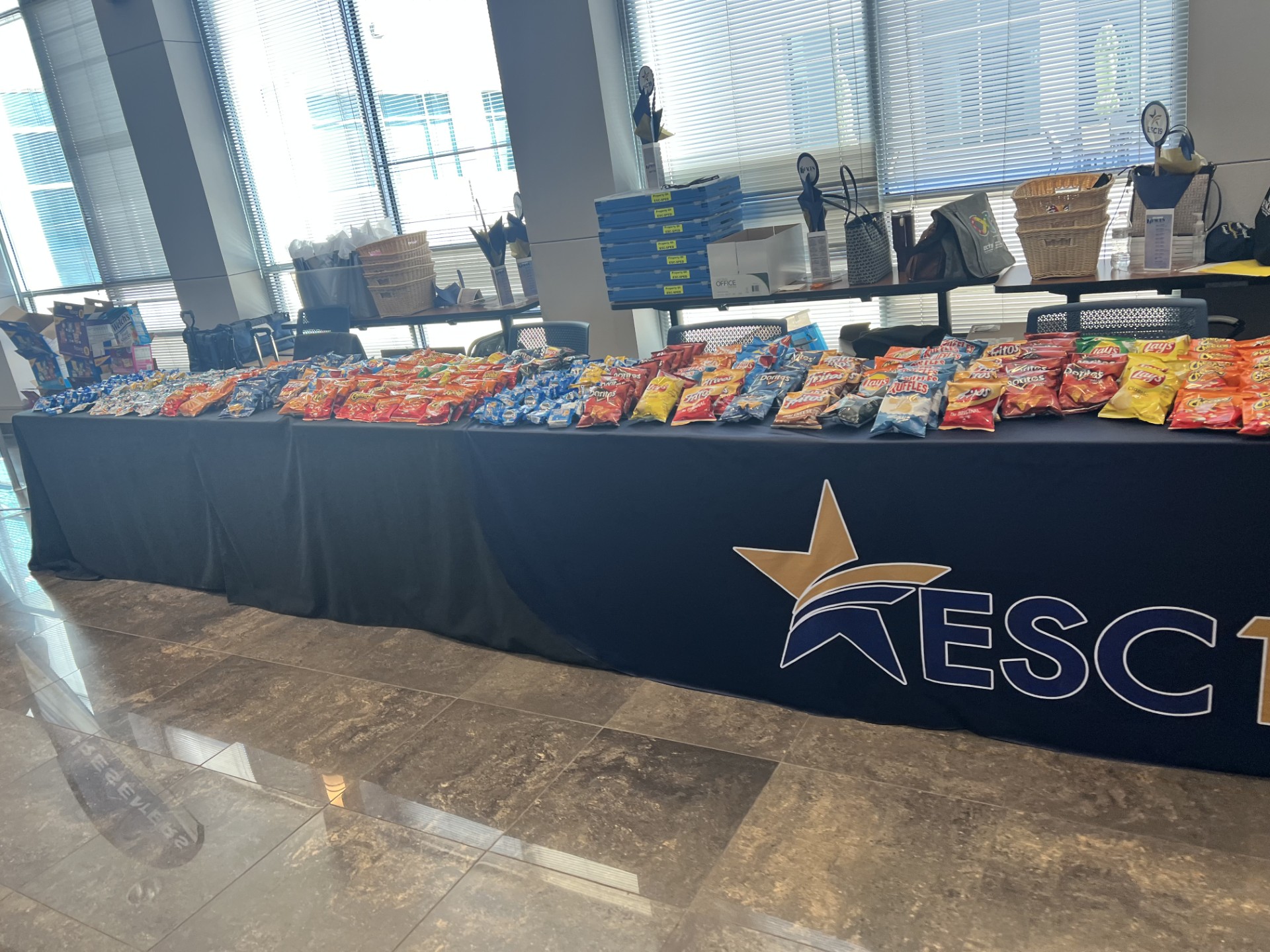 A long table laden with an assortment of snack bags is set up in front of a large windowed wall.