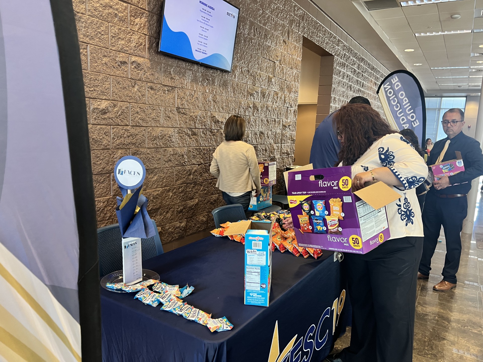 In the foreground, a woman unpacks boxes of snacks at a table draped with a blue cloth, while in the background, people mill about near a brick wall and a screen displaying information.