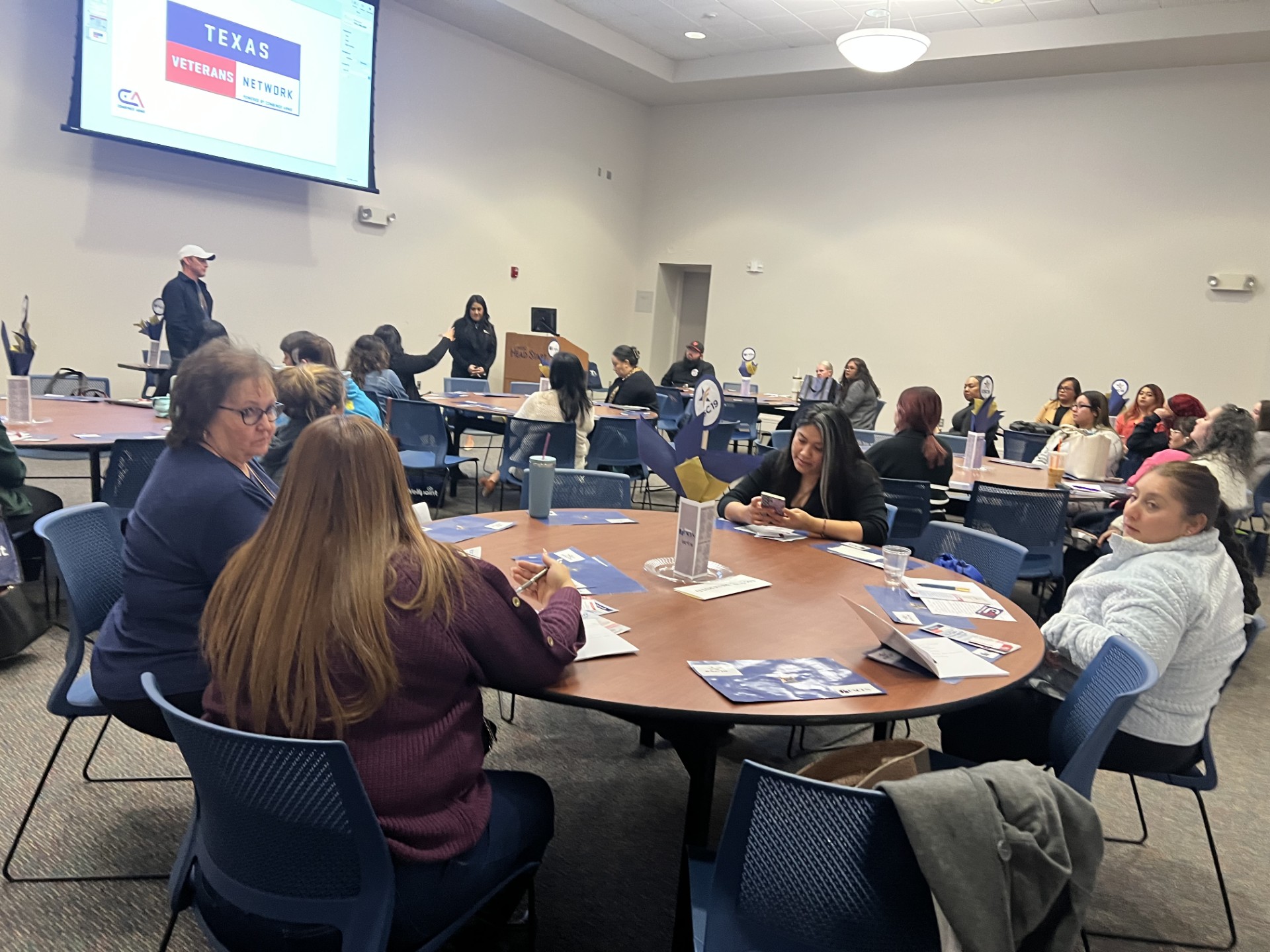 In the foreground, several women sit at a round table with papers and water bottles, while in the background, a man stands near a projector screen displaying "TEXAS VETERANS NETWORK."