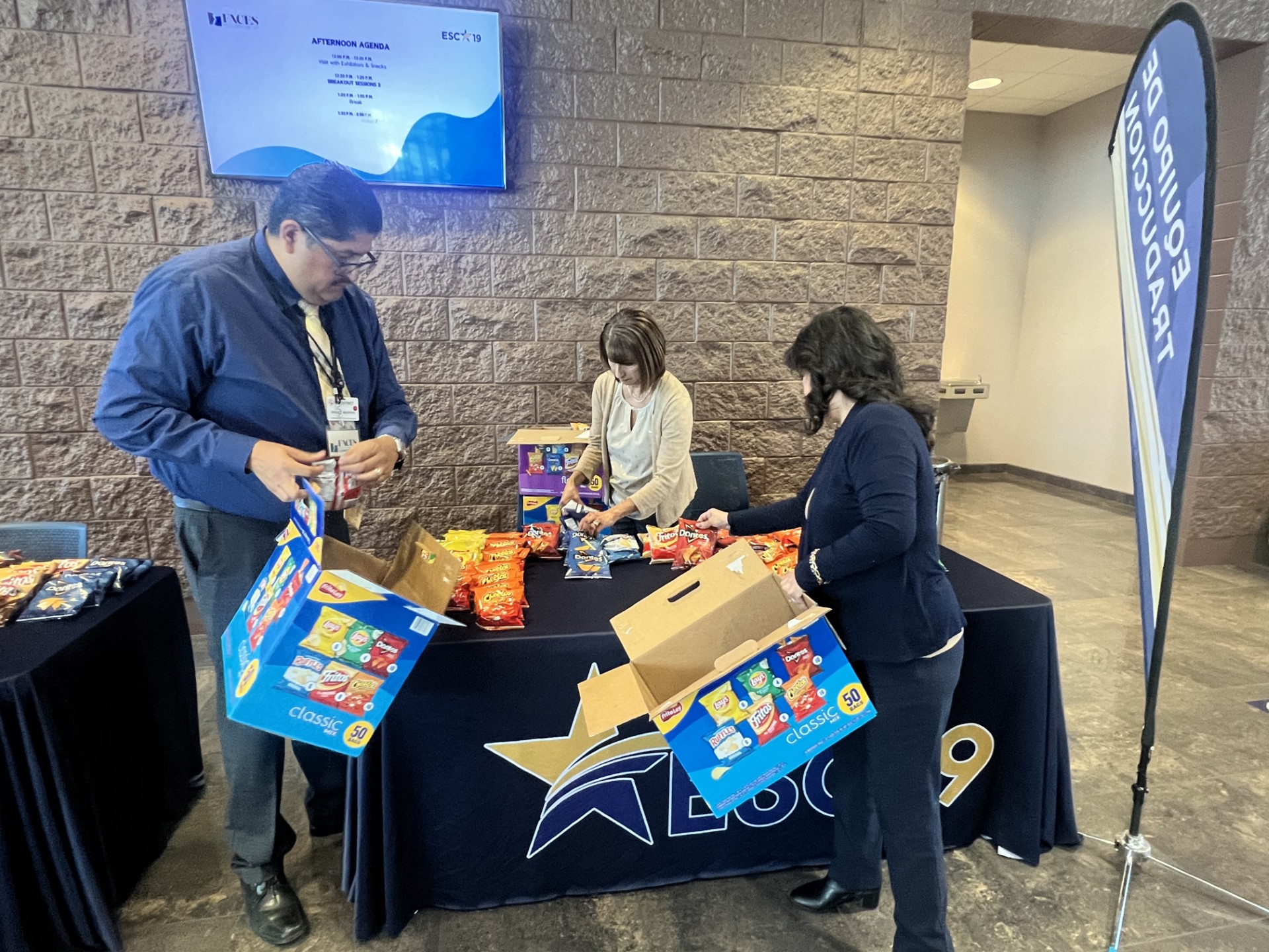 Three people are organizing snack boxes at a table with a brick wall and a screen in the background.