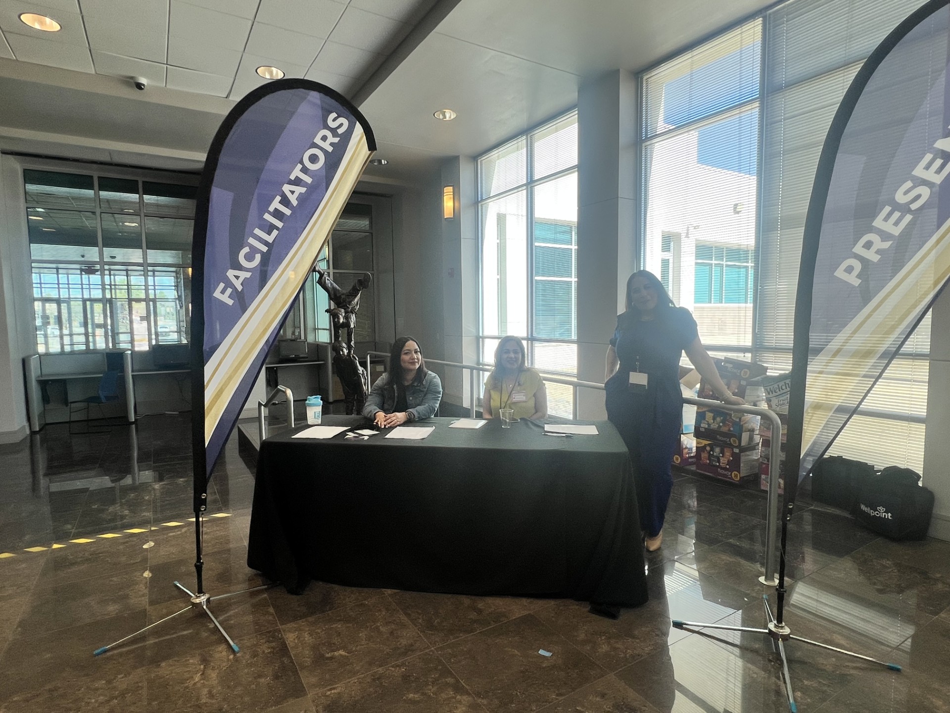 Two women sit behind a table with "FACILITATORS" and "PRESENTERS" flags in the foreground, while a bright, modern building with large windows forms the background.