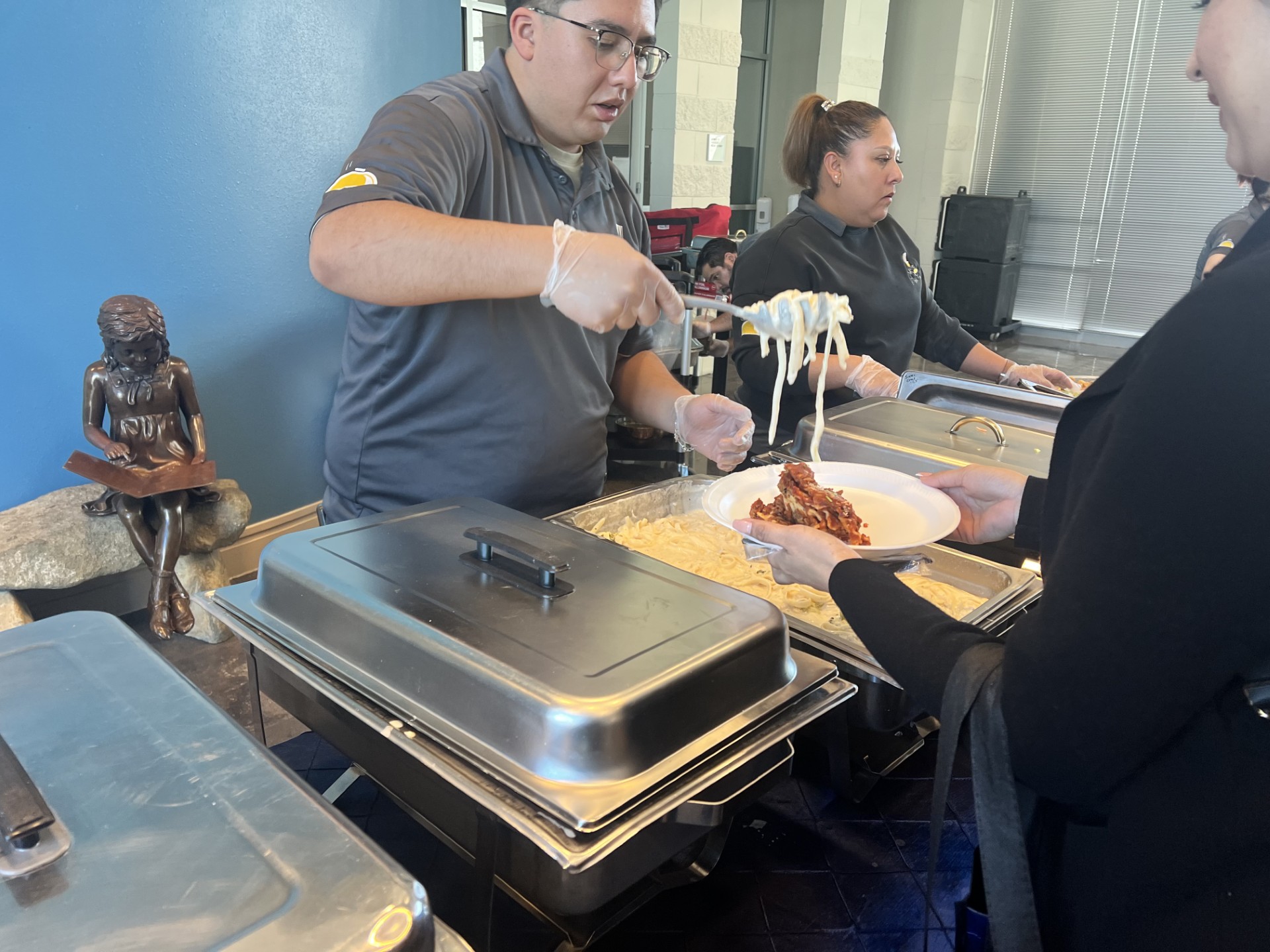 A man in a gray shirt serves pasta from a chafing dish to a woman in a black jacket, with a woman in a black shirt standing behind him and a small statue on a rock in the left foreground.