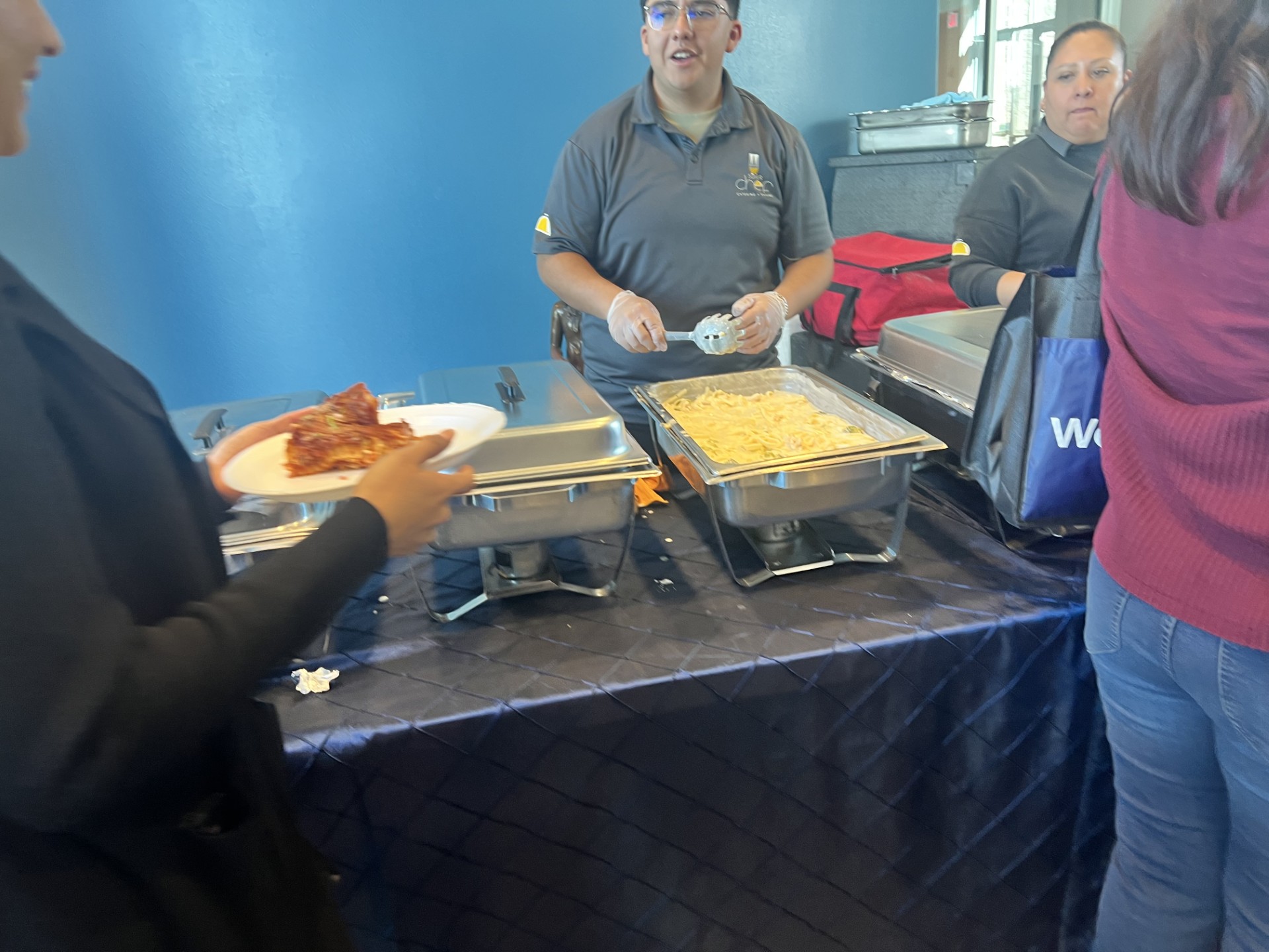 In the foreground, a person is served pizza from a buffet, while in the background, two servers stand behind chafing dishes filled with food.
