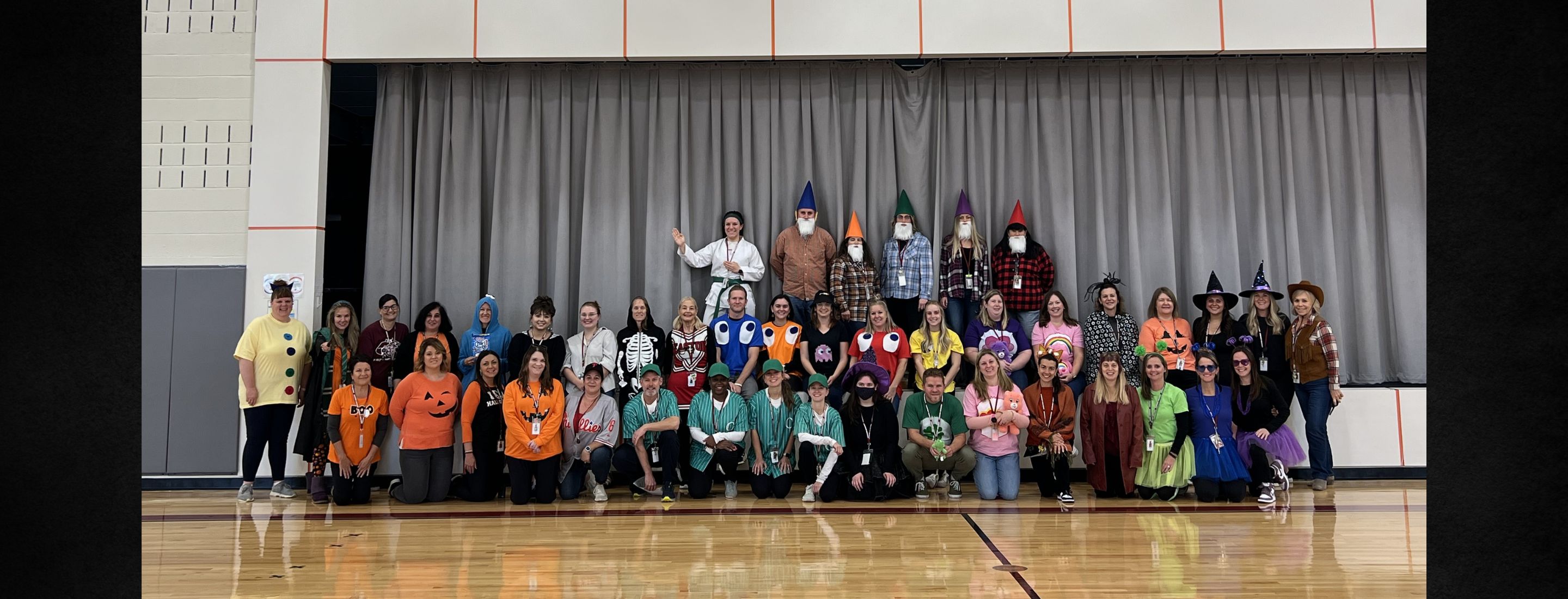 A group of adults are gathered for a photo in front of a stage with gray curtains closed behind them. The words Cheston Cougars appear on a white wall  above the stage. The adults are wearing various costumes.
