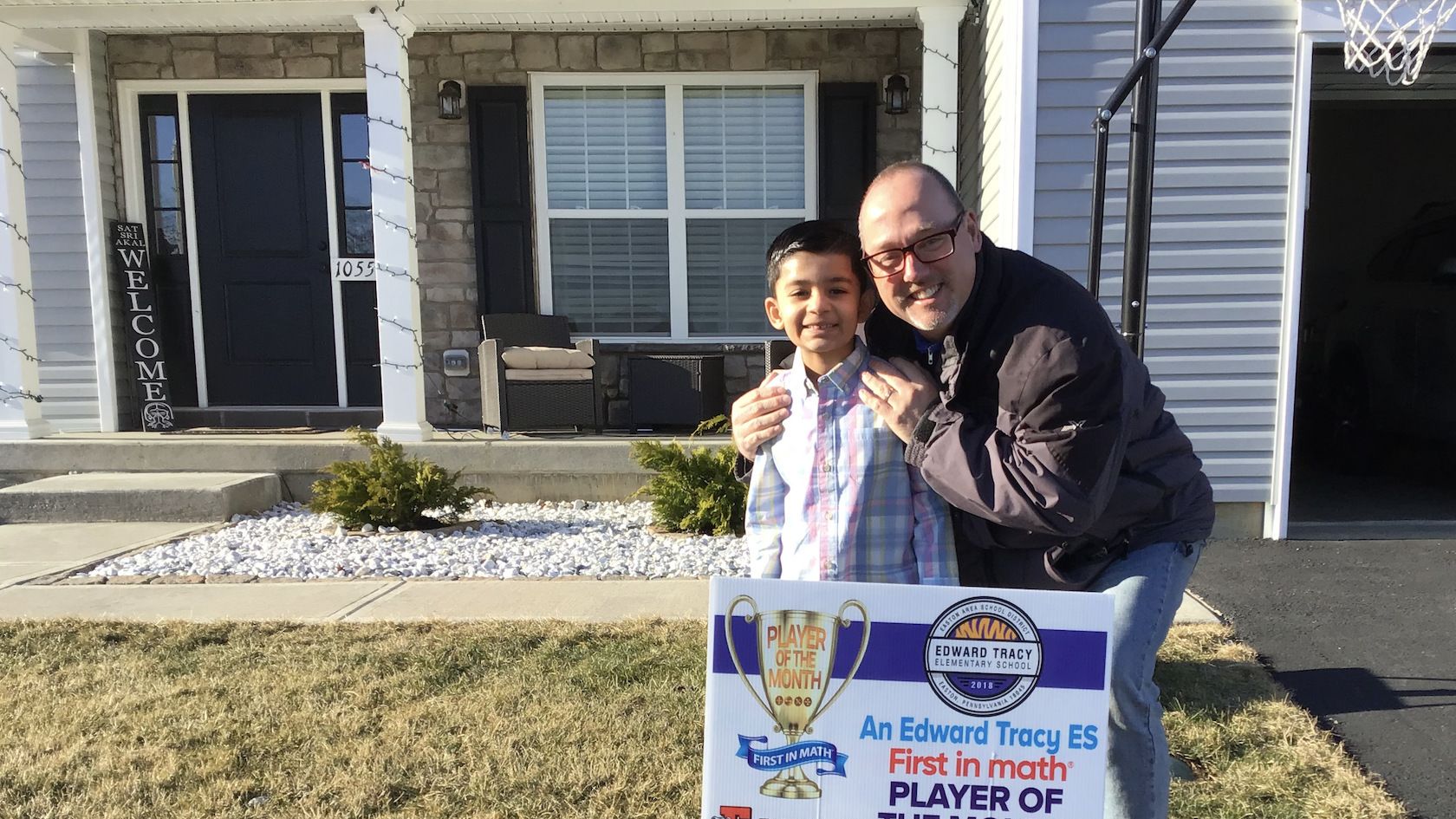 An elementary student and an adult male stand together on the lawn in front of a house. A yard sign congratulates the student on being a First in Math Player of the Month.