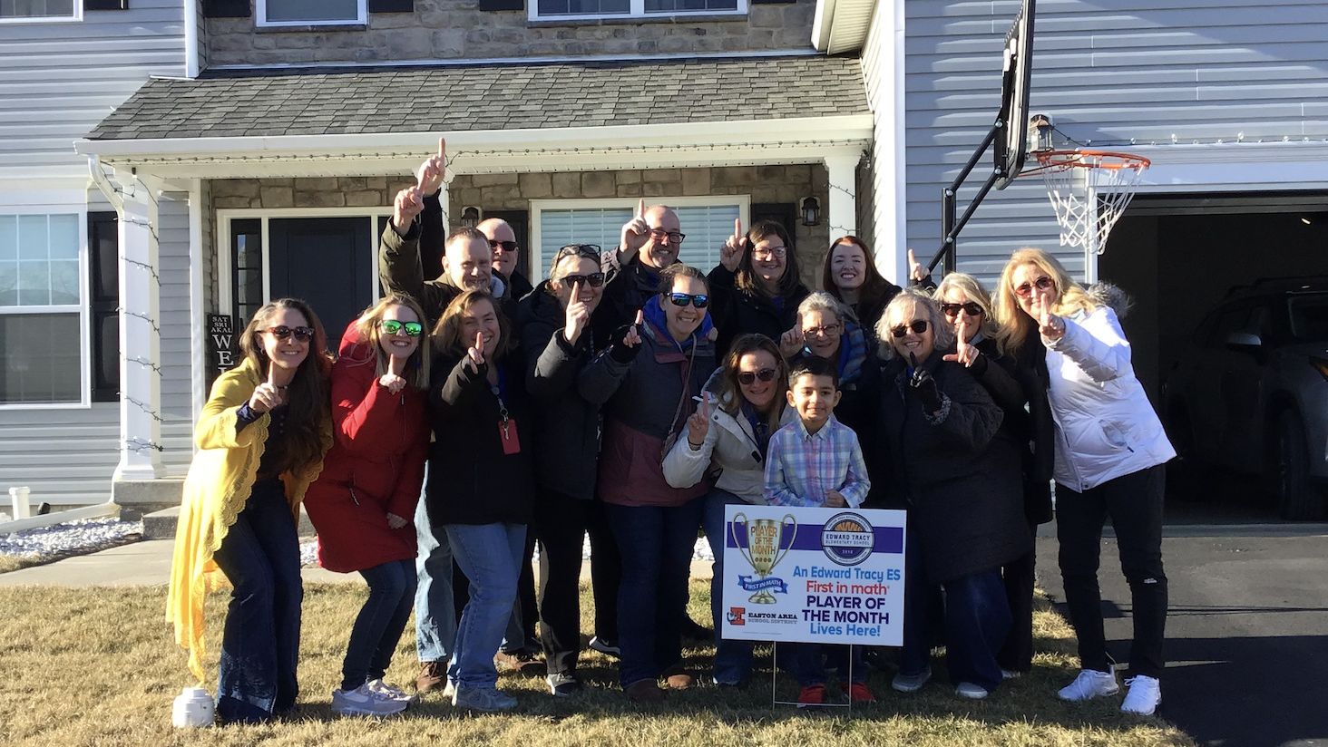 An elementary student and a group of adults stand together on the lawn in front of a house. A yard sign congratulates the student on being a First in Math Player of the Month.
