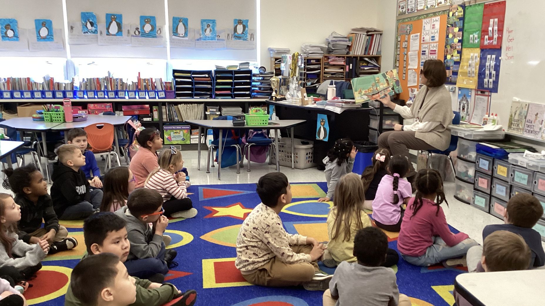 Elementary aged students sit on the floor in a classroom, on a colorful carpet, all looking at an adult sitting in a chair and reading a book aloud.