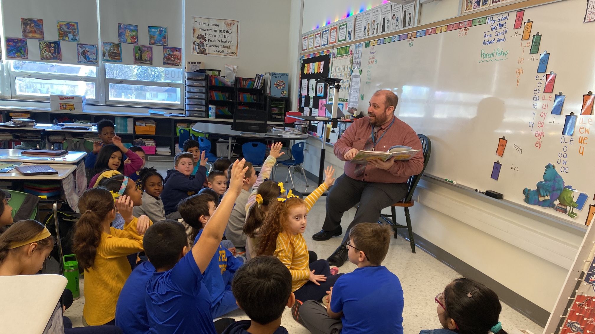 Elementary aged students sit on the floor in a classroom, on a colorful carpet, all looking at an adult sitting in a chair and reading a book aloud. A few students have their hands in the air.