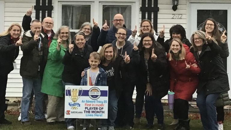 An elementary student and a group of adults stand together on the lawn in front of a house. A yard sign congratulates the student on being a First in Math Player of the Month.
