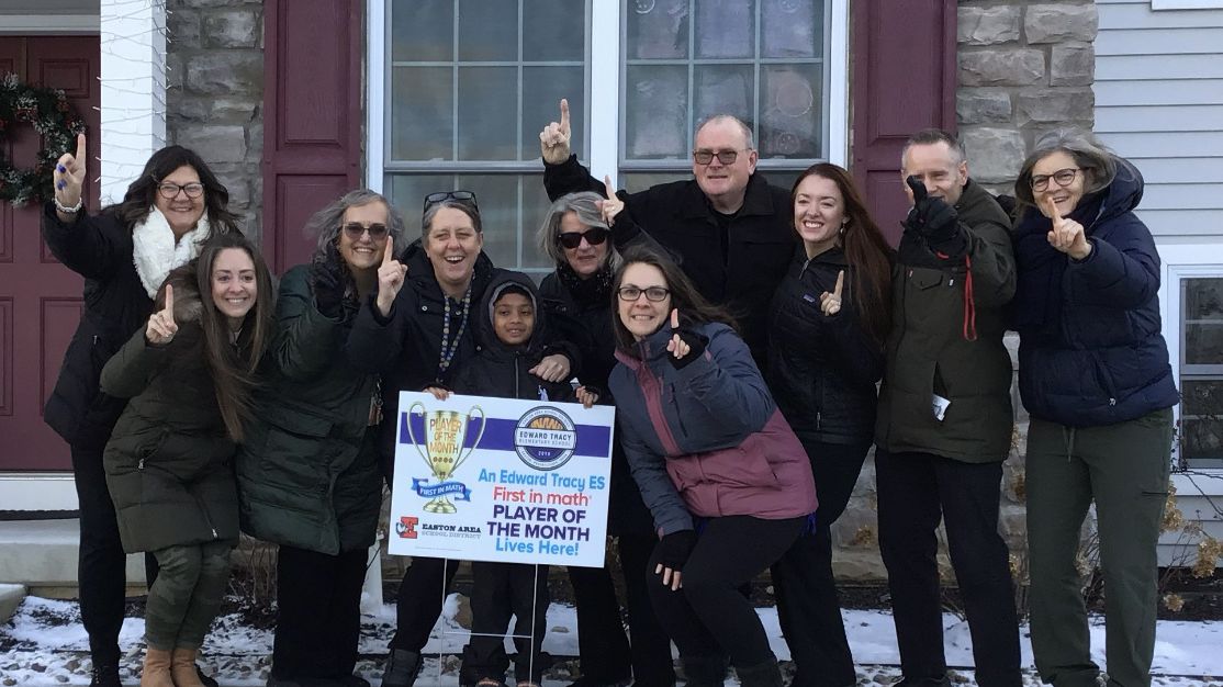 An elementary student and a group of adults stand together on the lawn in front of a house. A yard sign congratulates the student on being a First in Math Player of the Month.