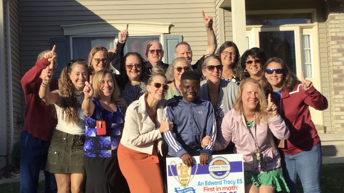 An elementary student and a group of adults stand together on the lawn in front of a house. A yard sign congratulates the student on being a First in Math Player of the Month.
