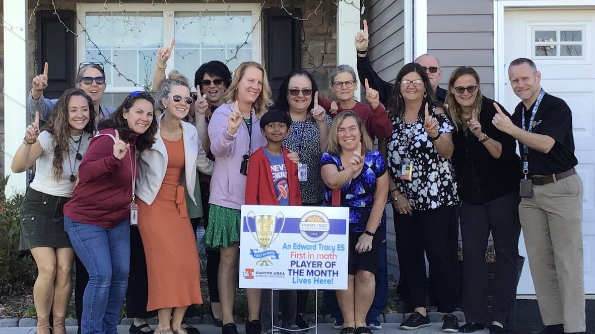 An elementary student and a group of adults stand together on the lawn in front of a house. A yard sign congratulates the student on being a First in Math Player of the Month.