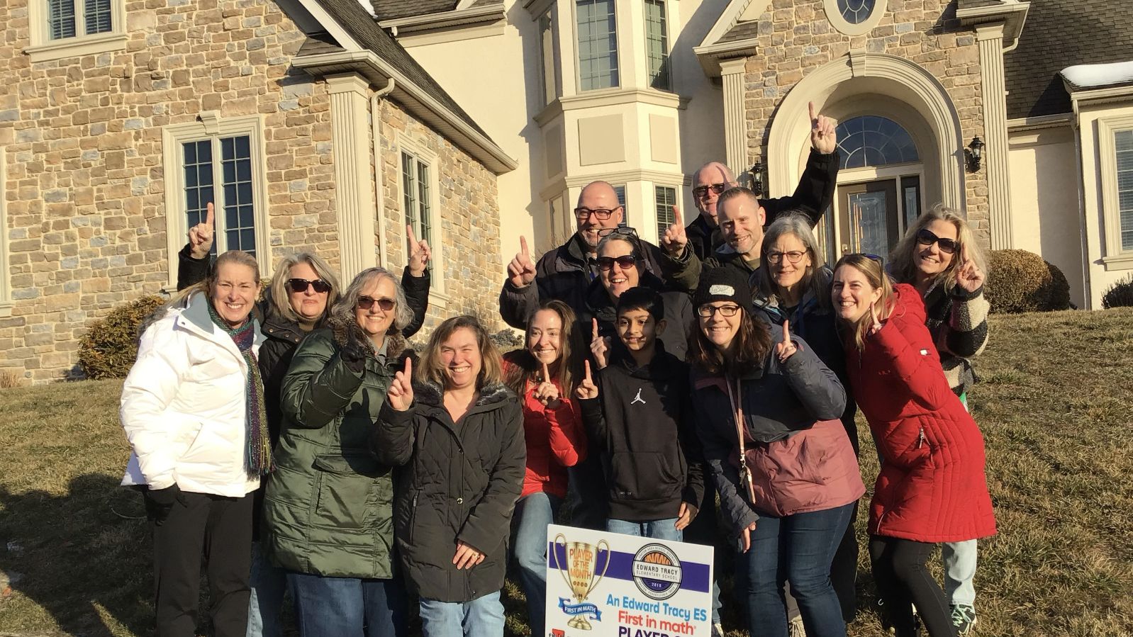 An elementary student and a group of adults stand together on the lawn in front of a house. A yard sign congratulates the student on being a First in Math Player of the Month.