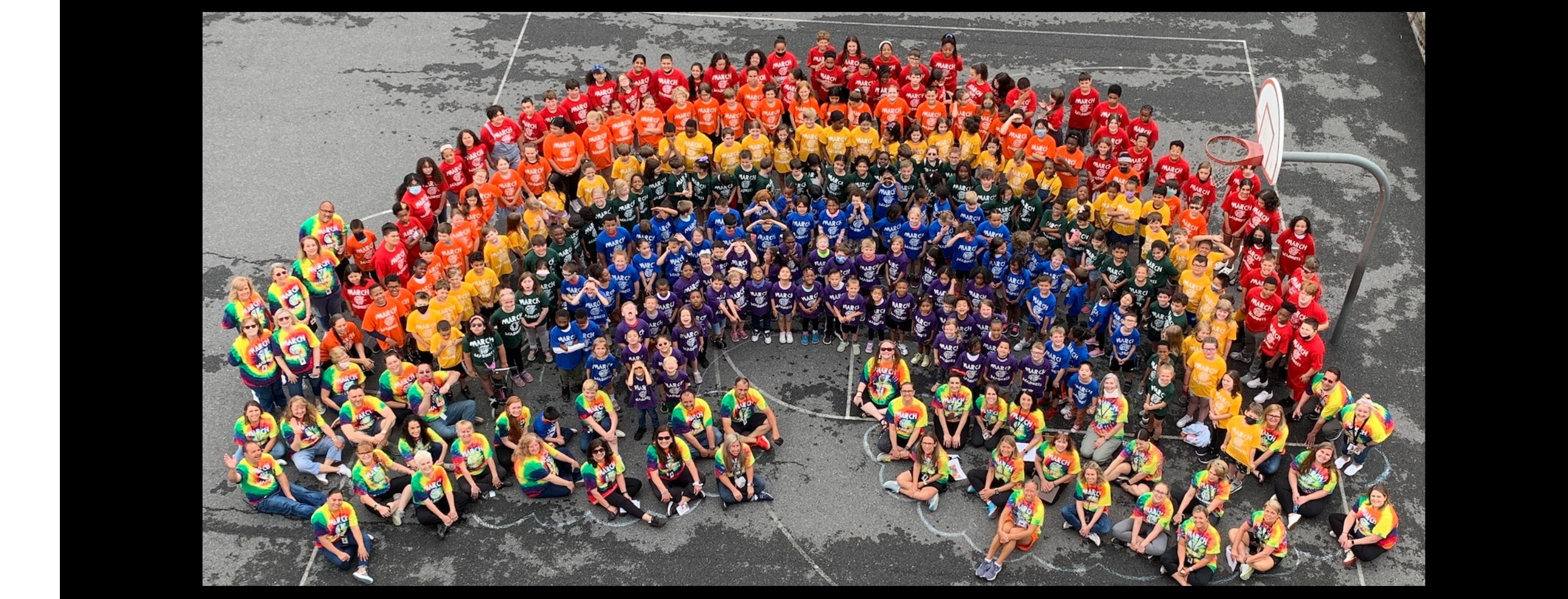An image take from above shows a large group of students and adults standing together to form a rainbow shape. In each row of the rainbow, students wear the same color t-shirt so that the outer line appears red, next orange, then yellow, green, blue, and purple. Adults wear multi-colored tie-dyed shirts and surround the ends of the rows to give the appearance of clouds.