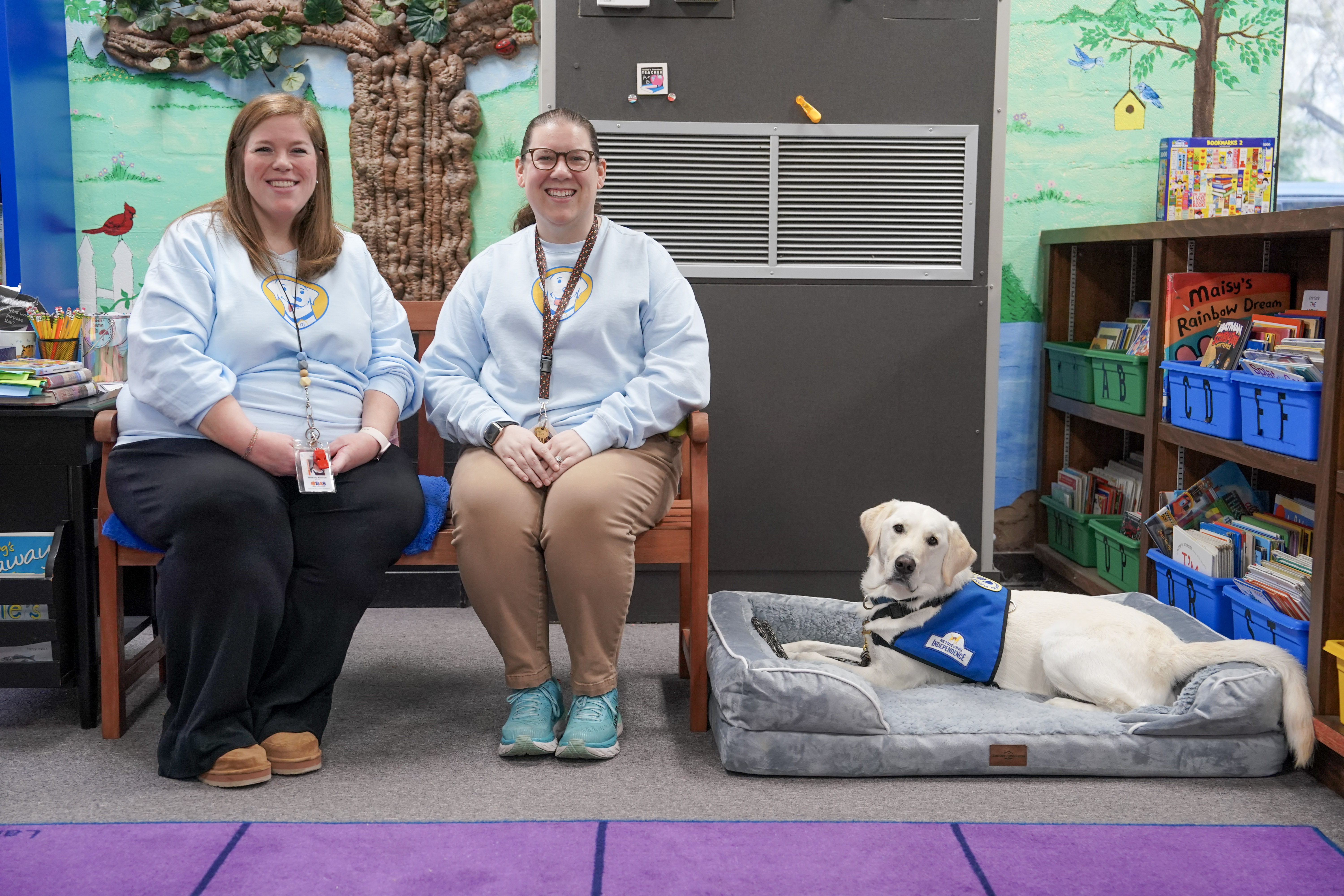 facility dog in a bed on the floor of library with two women seated on bench