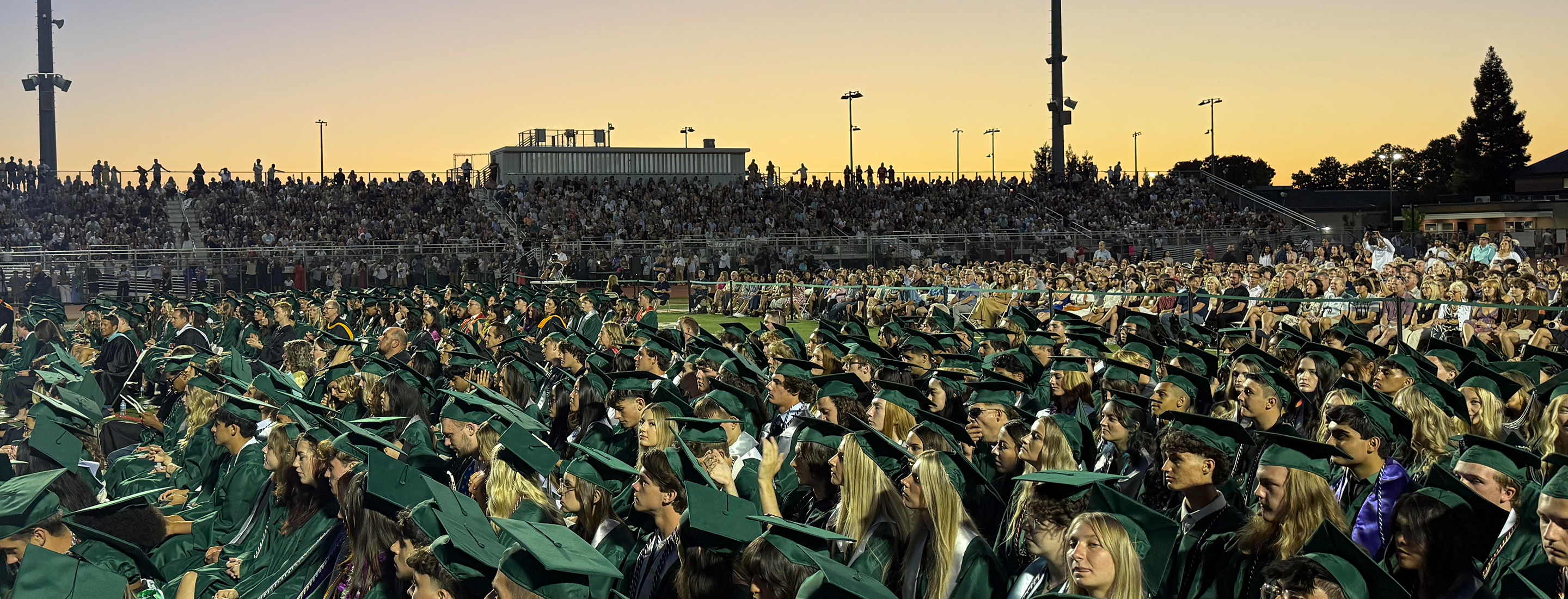 A large crowd of people in green graduation gowns and caps seated in a stadium, with a building and lights visible in the background.