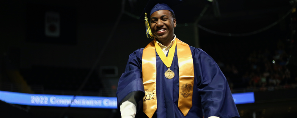 A young man in a blue graduation gown and yellow sash stands on a stage, holding a medal around his neck and smiling.