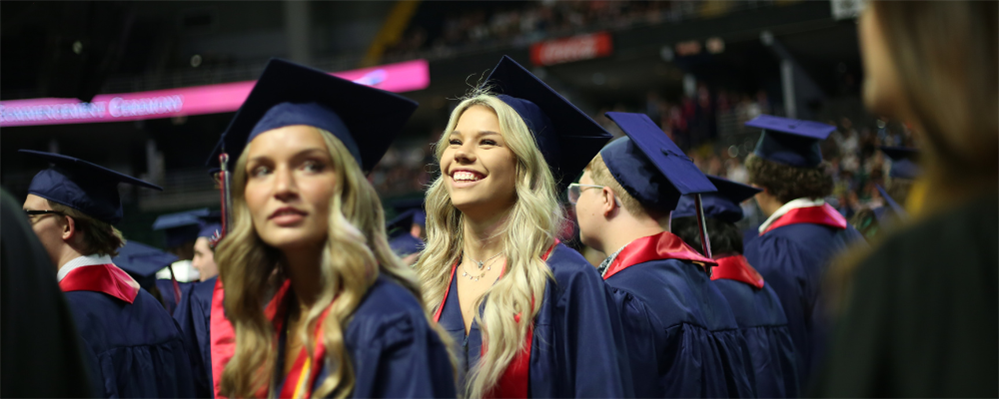 The image shows a group of smiling graduates in graduation gowns and caps, standing together in what appears to be a ceremony or event.