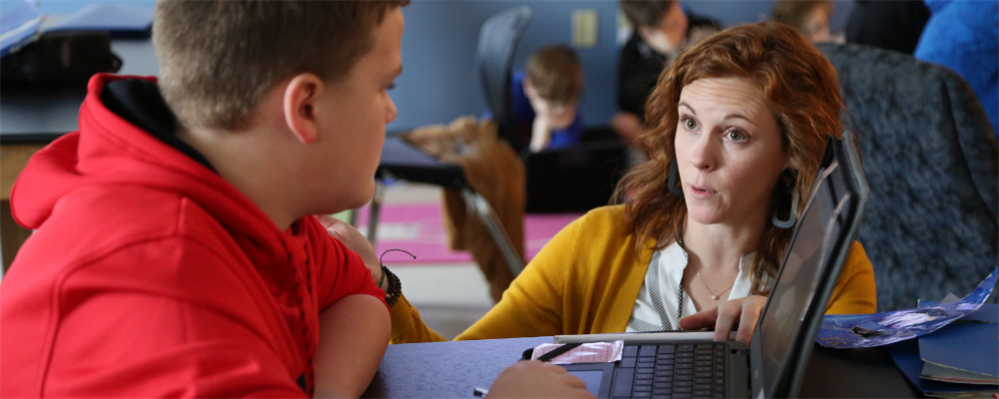Two people, a young boy in a red shirt and a woman with reddish-brown hair in a yellow sweater, are engaged in a conversation while the woman appears to be using a laptop computer in what seems to be a classroom or educational setting.