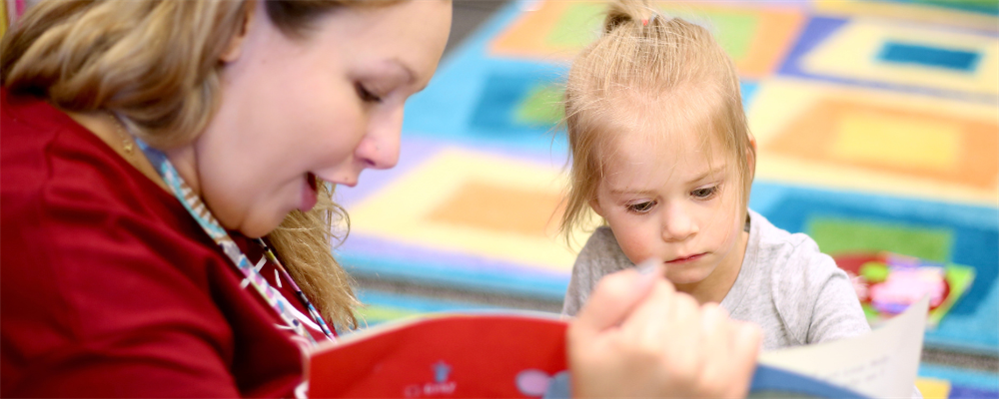 A young woman in a red shirt is reading a book with a young child sitting next to her, surrounded by colorful artwork on the walls.