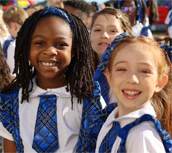 The image shows three young girls in school uniforms, smiling and standing together. They appear to be of different ethnic backgrounds, with one girl having dreadlocks and the other two having lighter hair. The background suggests they are in a school setting, with other students visible in the background.