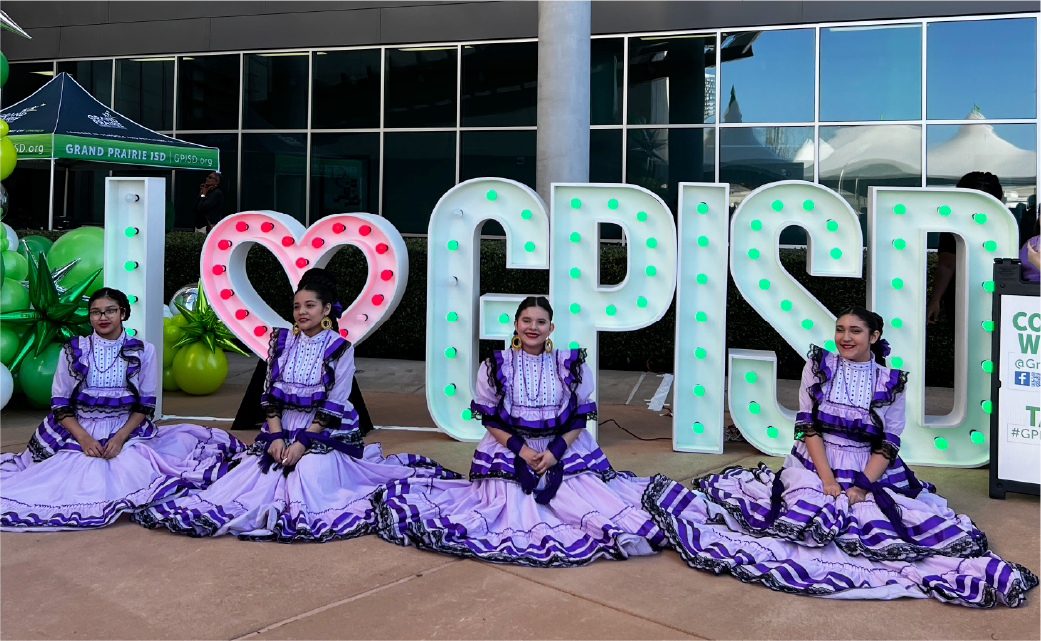 The image shows a group of people dressed in elaborate purple and white costumes, posing in front of a large "I Love Corpus" sign against a backdrop of tents and other festival-like structures.