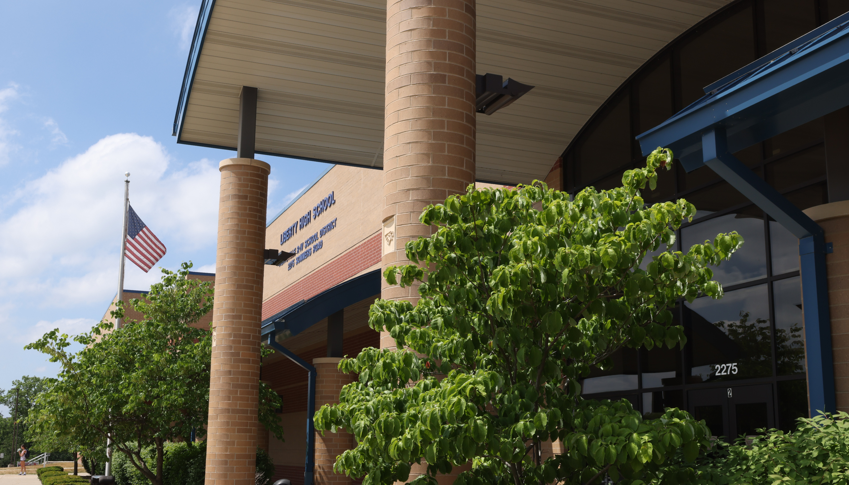 A brick building with a covered porch, an American flag, and a tree with lush green foliage in the foreground against a clear blue sky.