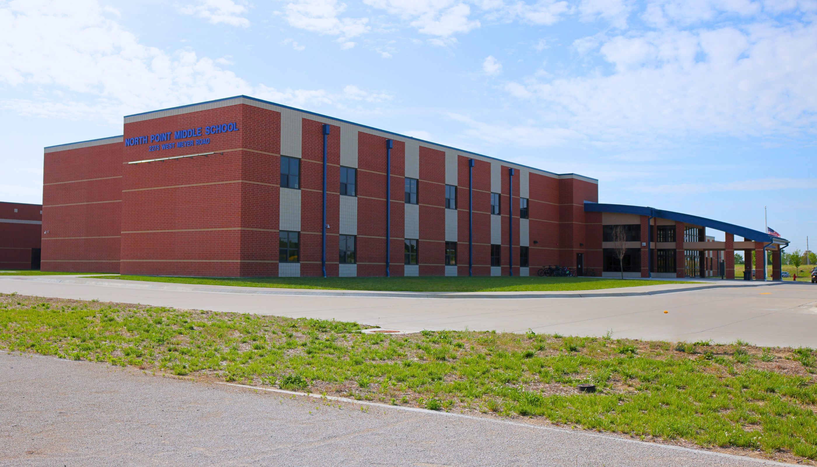 A large, multi-story brick building with a curved entrance and a grassy area in the foreground, set against a blue sky with scattered clouds.