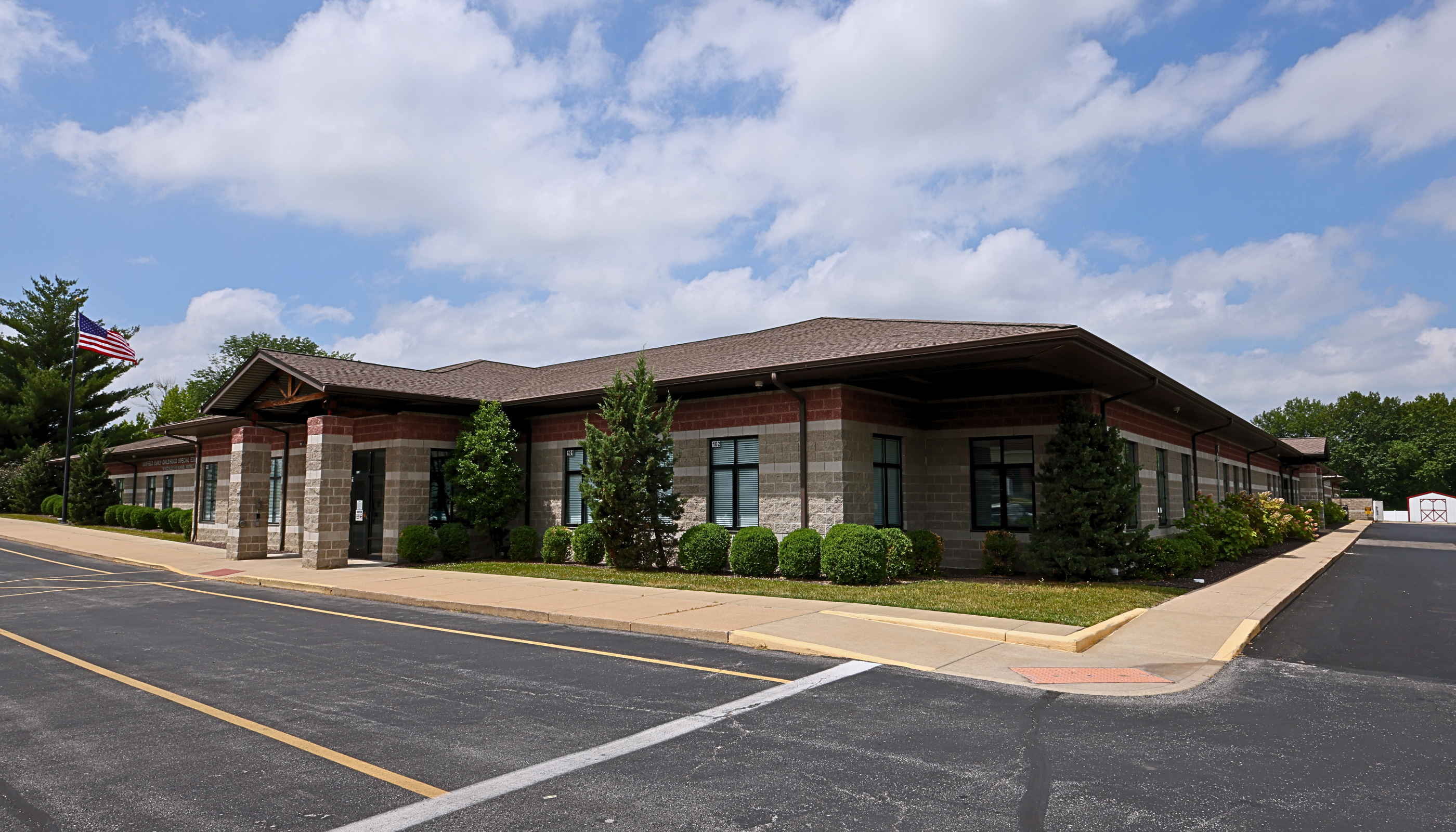 A modern, multi-story building with a brick exterior and a sloped roof stands in the foreground, surrounded by a paved parking lot and lush greenery in the background against a cloudy blue sky.