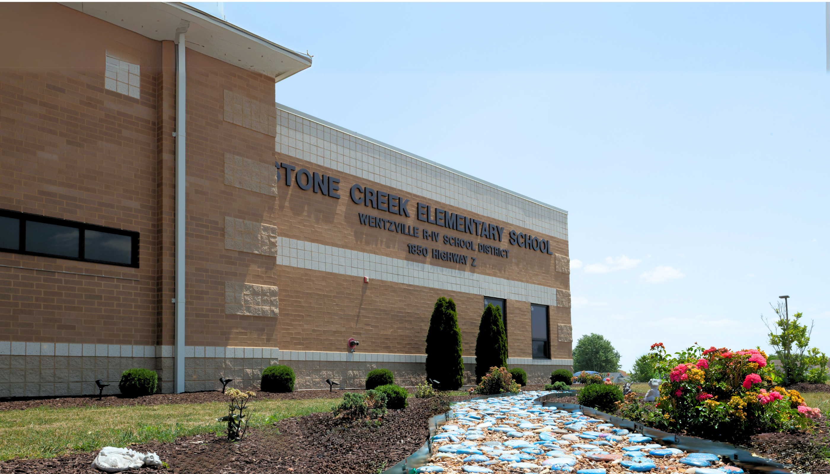 A brick building with the name "Lone Creek Elementary School" displayed prominently, surrounded by a landscaped area with a decorative stream and flowering plants in the foreground.