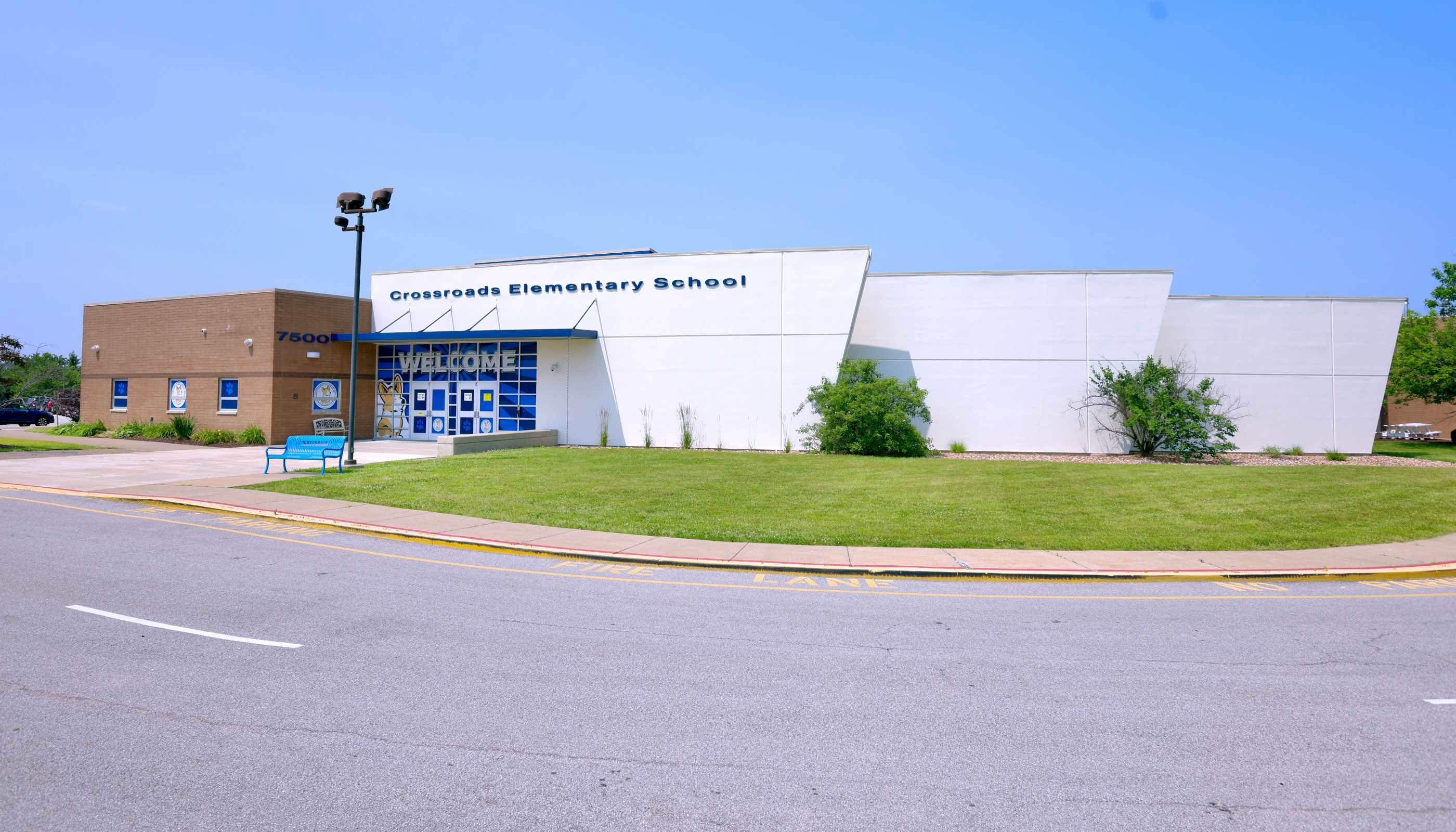 A modern, single-story elementary school building with a grassy lawn and a paved road in the foreground.