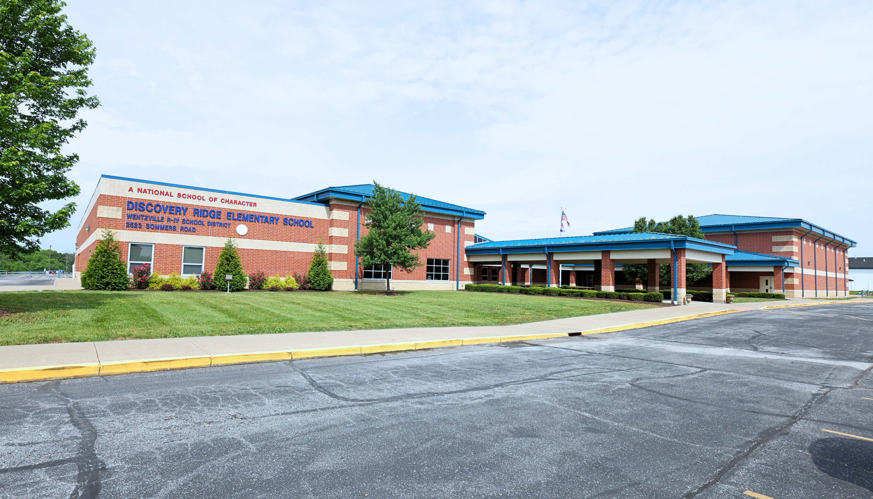 A large brick building with a blue roof and a covered entrance area, surrounded by trees and a paved parking lot in the foreground.