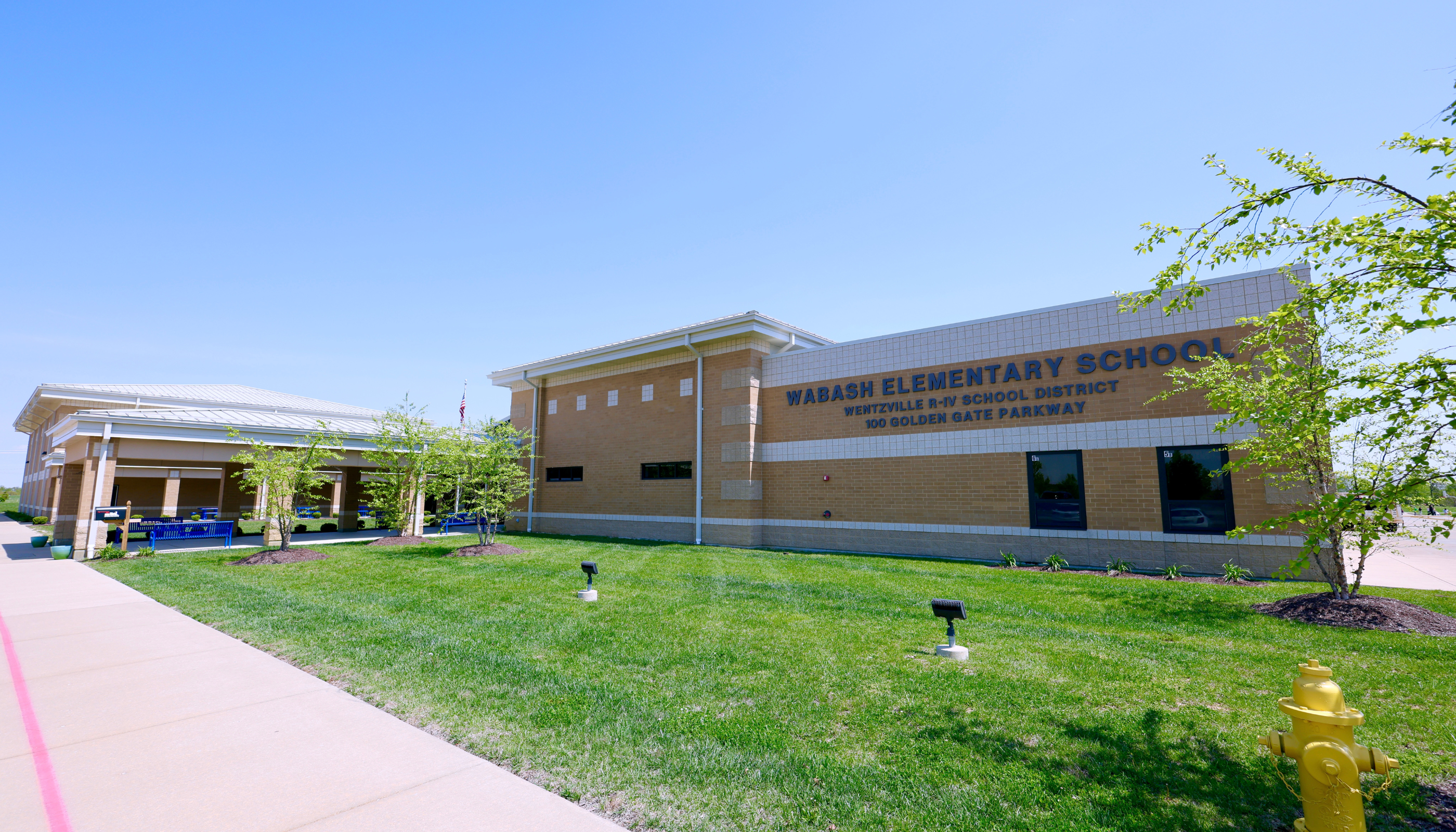 A modern, two-story elementary school building with a grassy lawn and landscaping in the foreground, set against a clear blue sky.