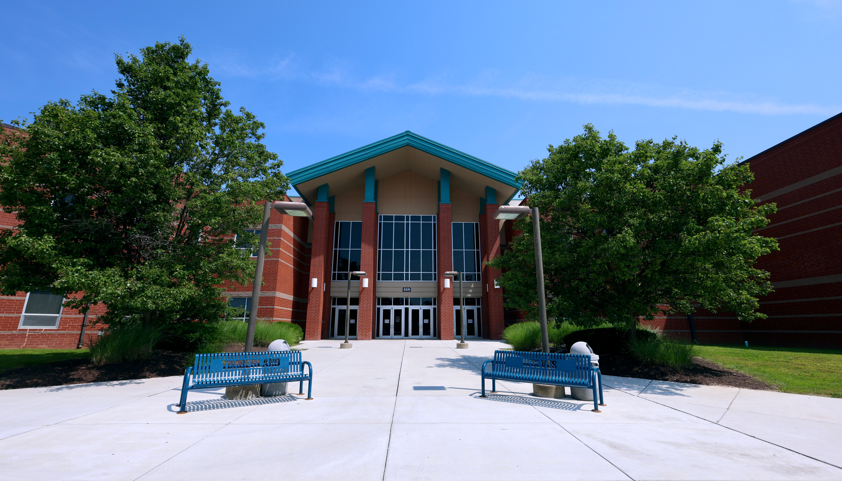 A modern brick building with a green roof and large windows stands in the background, surrounded by lush green trees. In the foreground, two blue benches are placed on a concrete walkway leading up to the building's entrance.