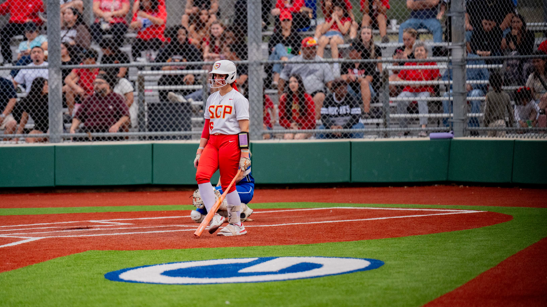 A baseball player in a red uniform stands on the pitcher's mound, surrounded by a crowd of spectators in the background.