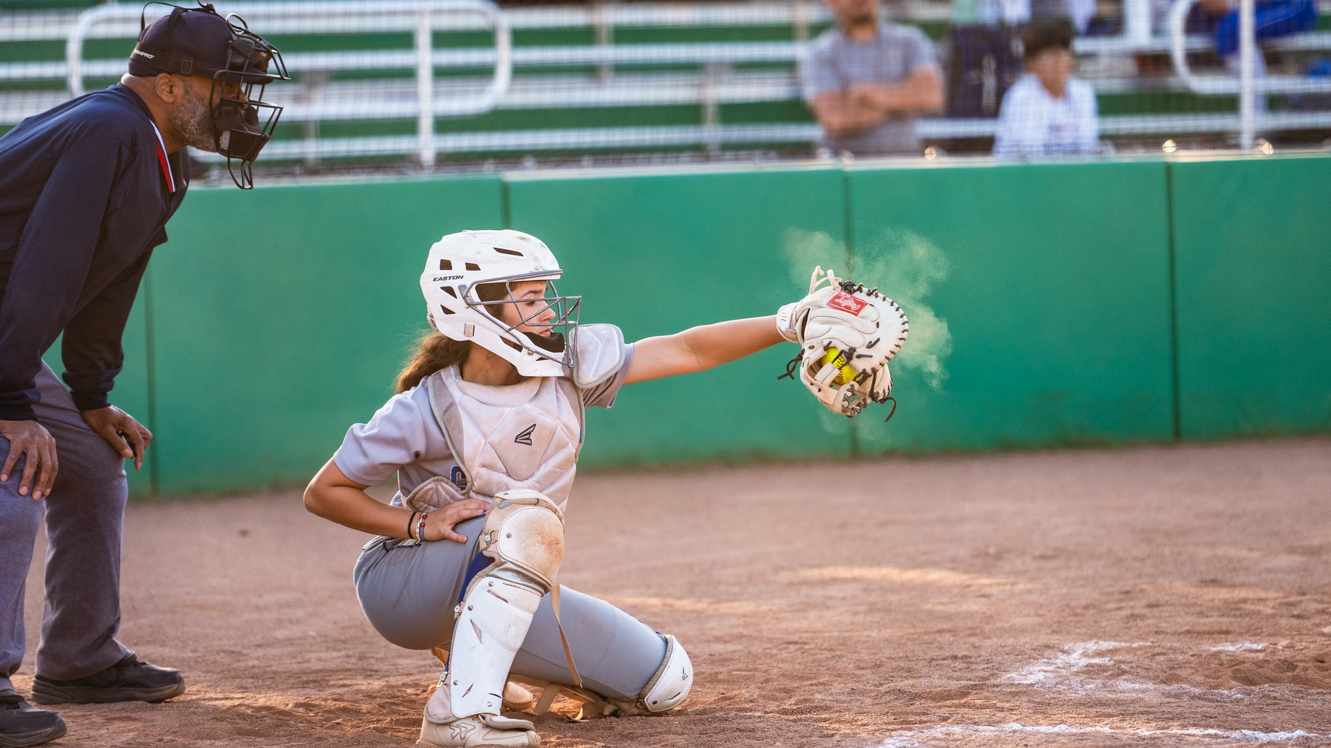 A young female softball player in full gear is kneeling on the field, while an umpire stands nearby on the baseball diamond surrounded by a green outfield fence.