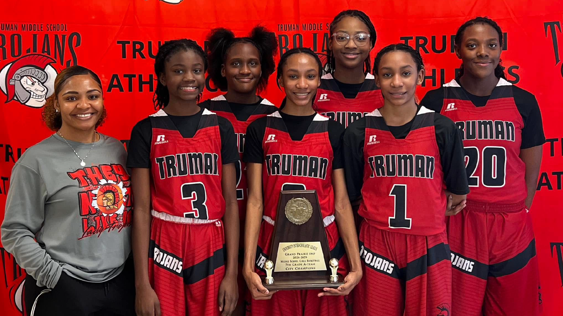 A group of young basketball players, wearing red and black uniforms, stand together holding a trophy in front of a red background with the word "Truman" prominently displayed.