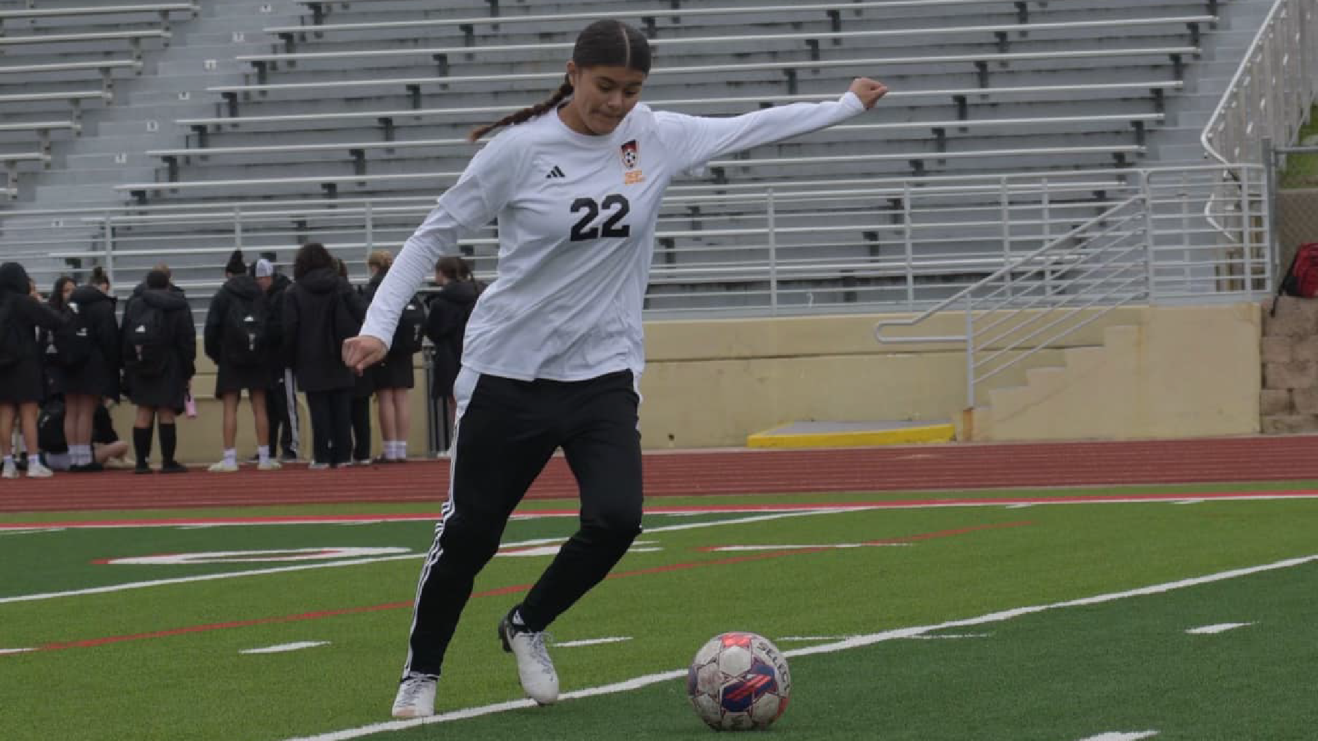 A soccer player in a white jersey stands on a field, with a group of people in the background.