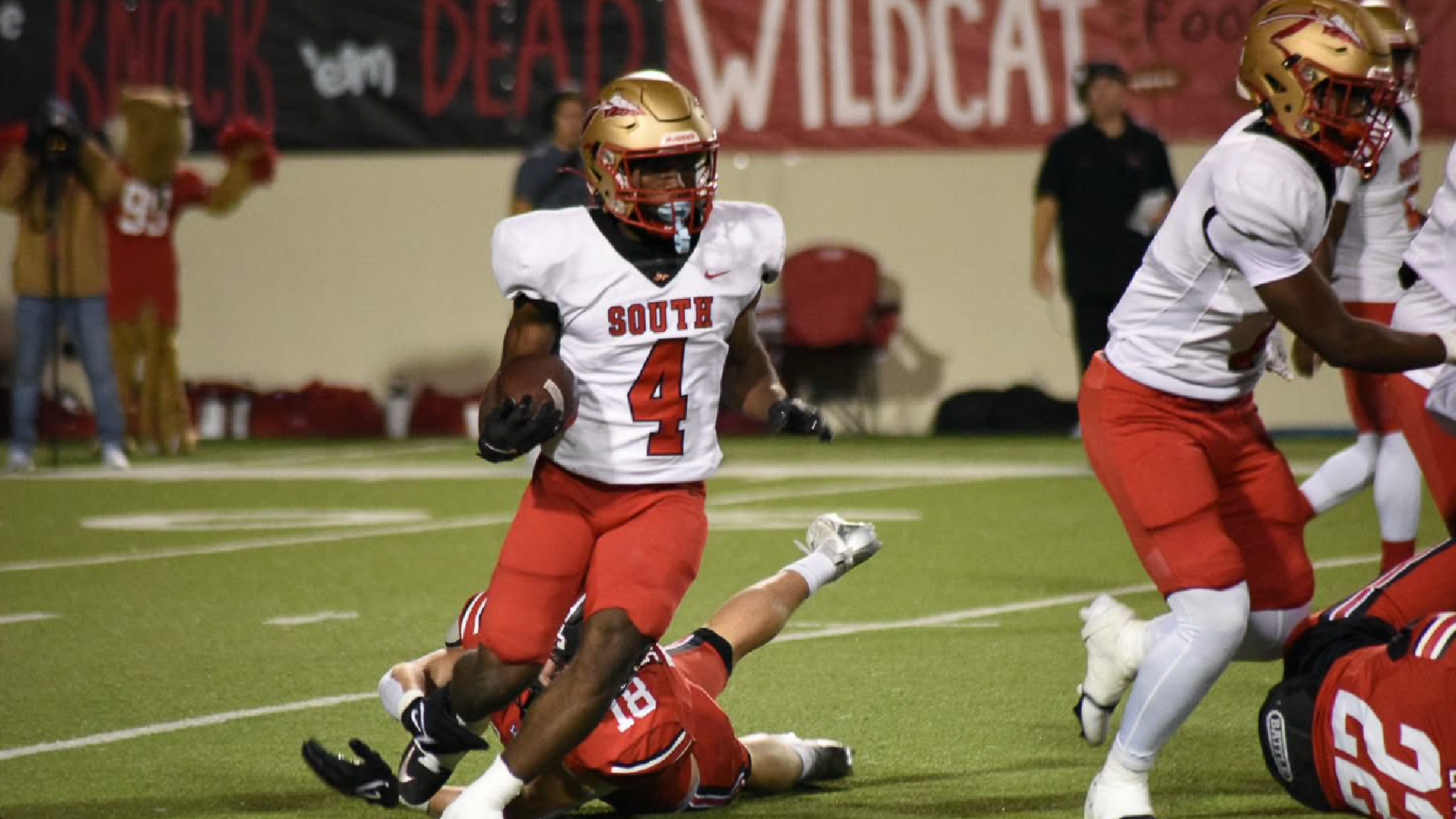The image shows two football players in uniform, one lying on the ground and the other standing over them on a football field with a red and white backdrop.
