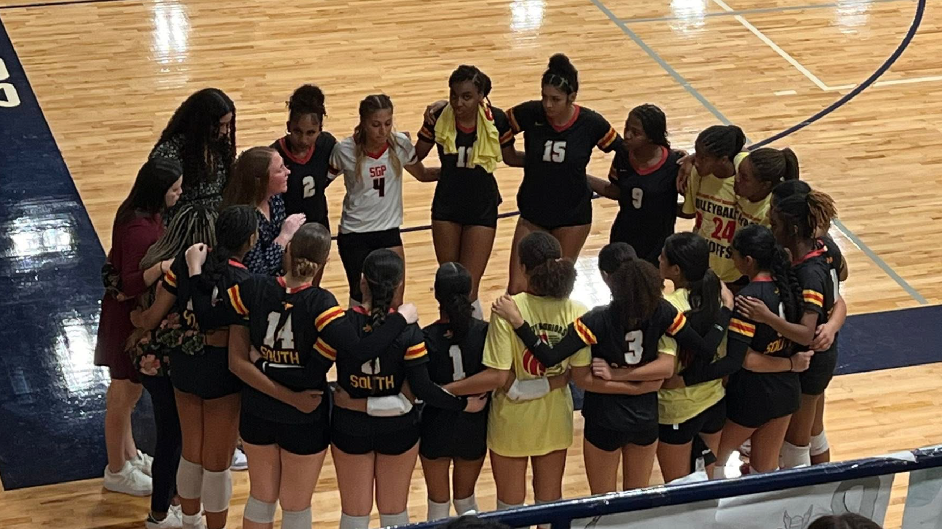 A group of young female athletes, likely a volleyball team, gathered together on a hardwood basketball court, with a scoreboard visible in the background.