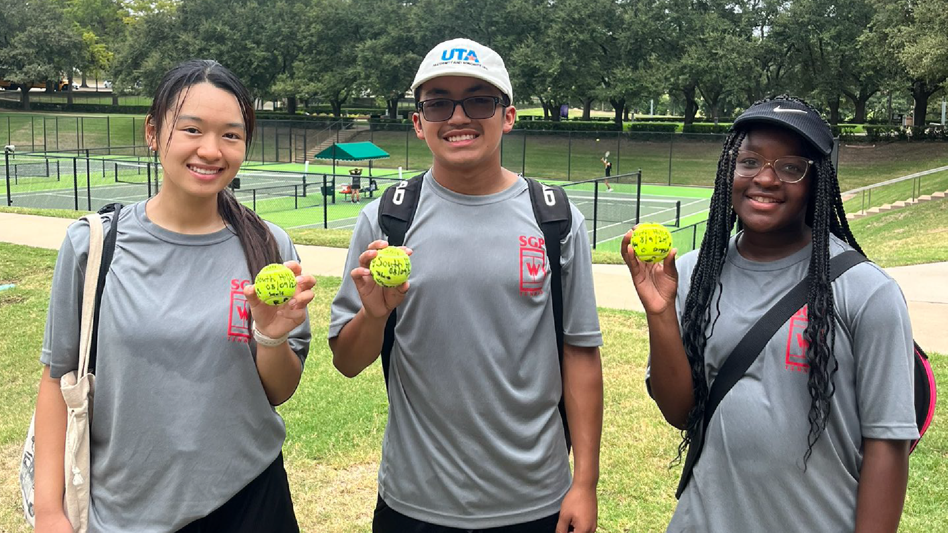 Three young people, two women and one man, are standing on a grassy field holding tennis balls and wearing athletic clothing. In the background, there are trees and what appears to be a tennis court.