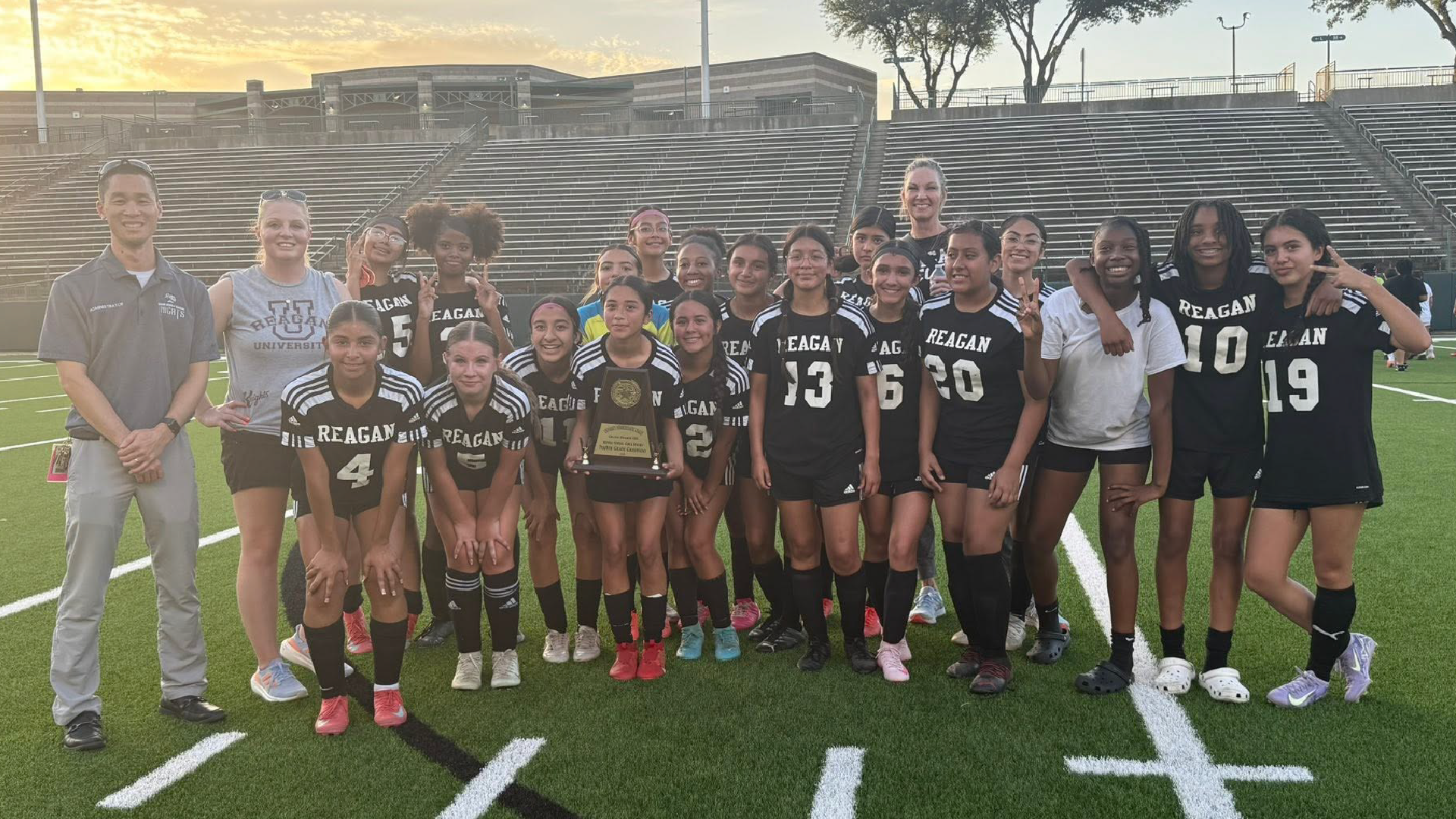 A group of young female soccer players posing together on a soccer field, with a stadium-like structure visible in the background.