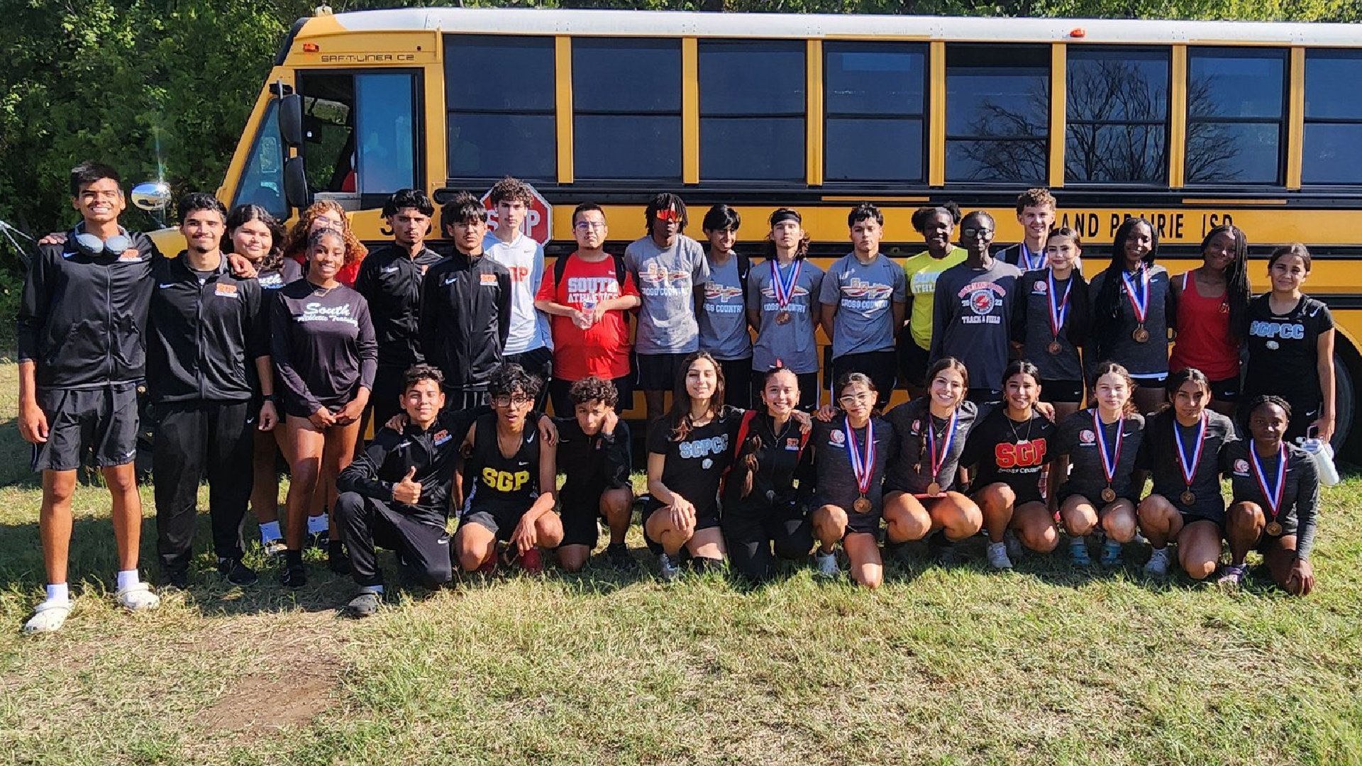 A group of young people, likely a sports team or club, posing together in front of a yellow school bus on a grassy field with trees in the background.