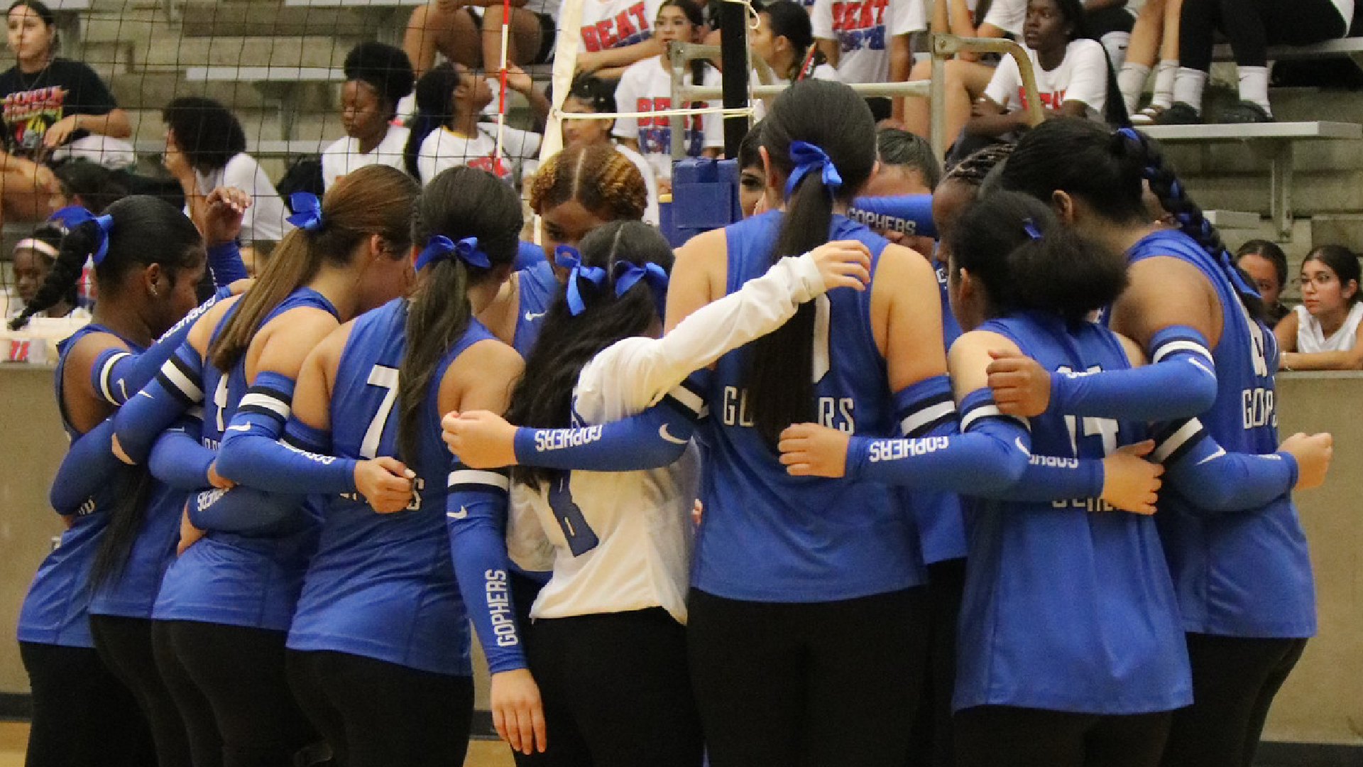 A group of young female athletes in blue uniforms standing together, with a crowd of spectators visible in the background.