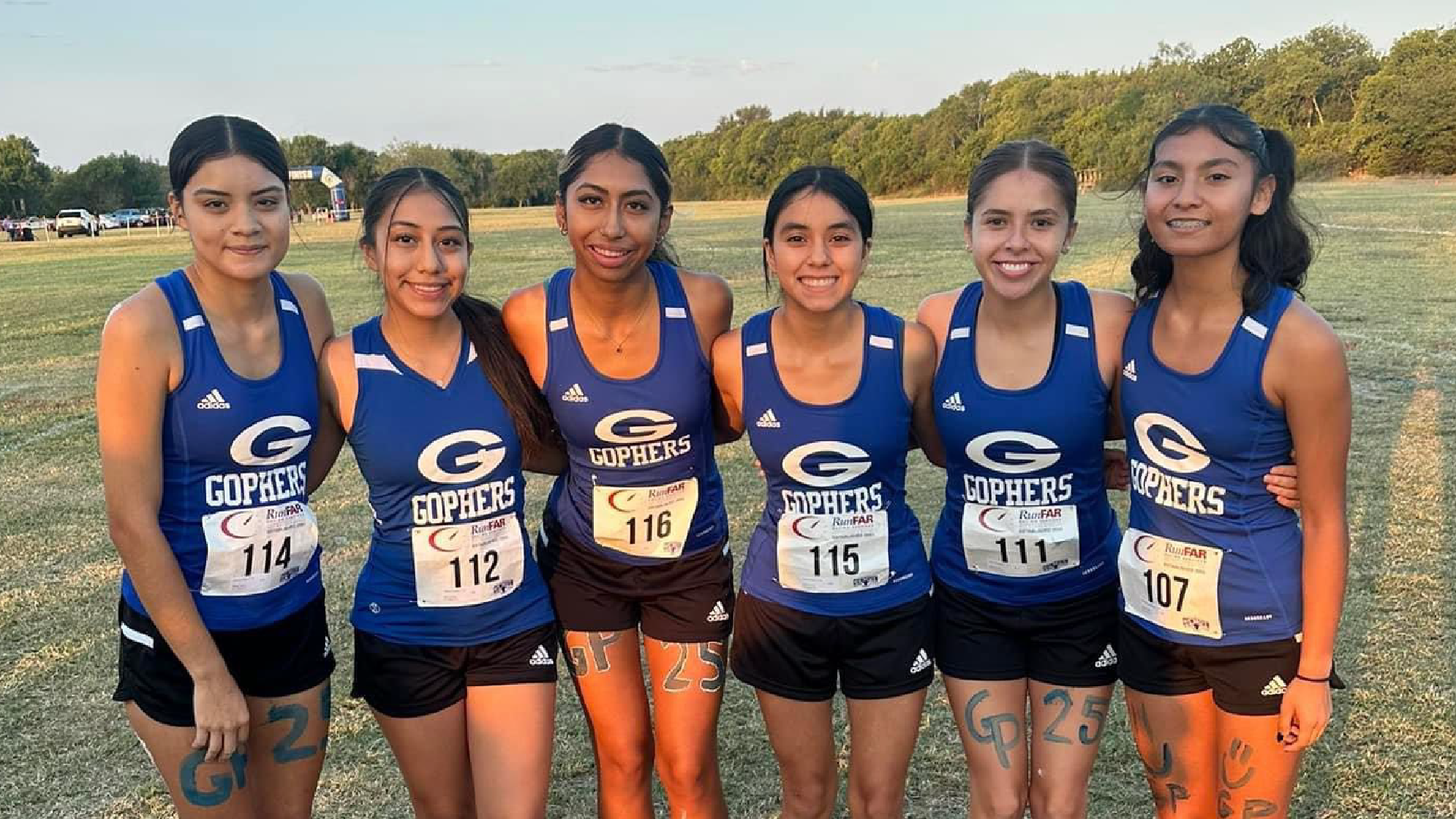 A group of six young women in athletic uniforms stand together on a grassy field, with a forested area visible in the background.