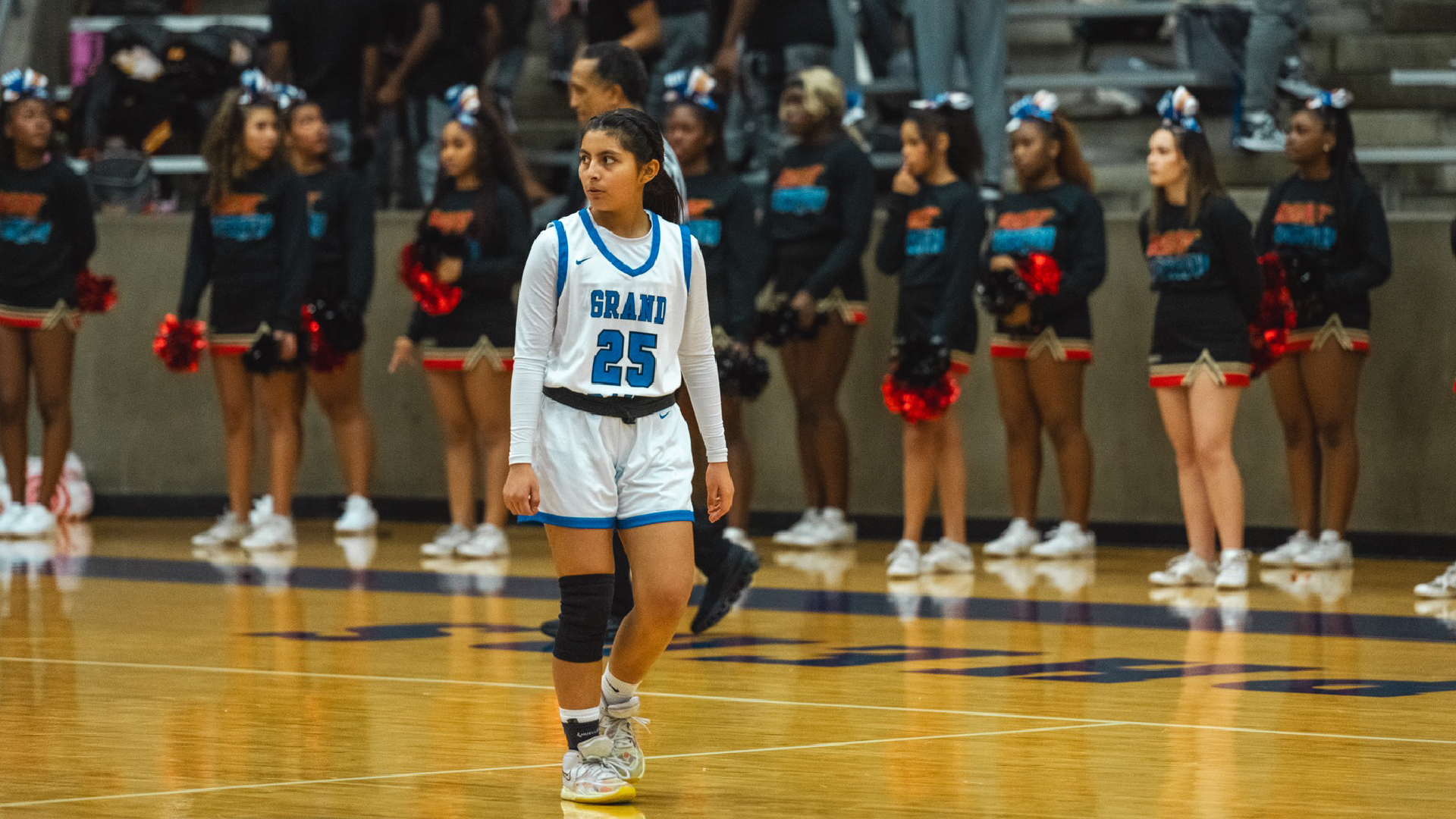 A young woman in a basketball jersey stands on a basketball court, surrounded by a group of cheerleaders in the background.