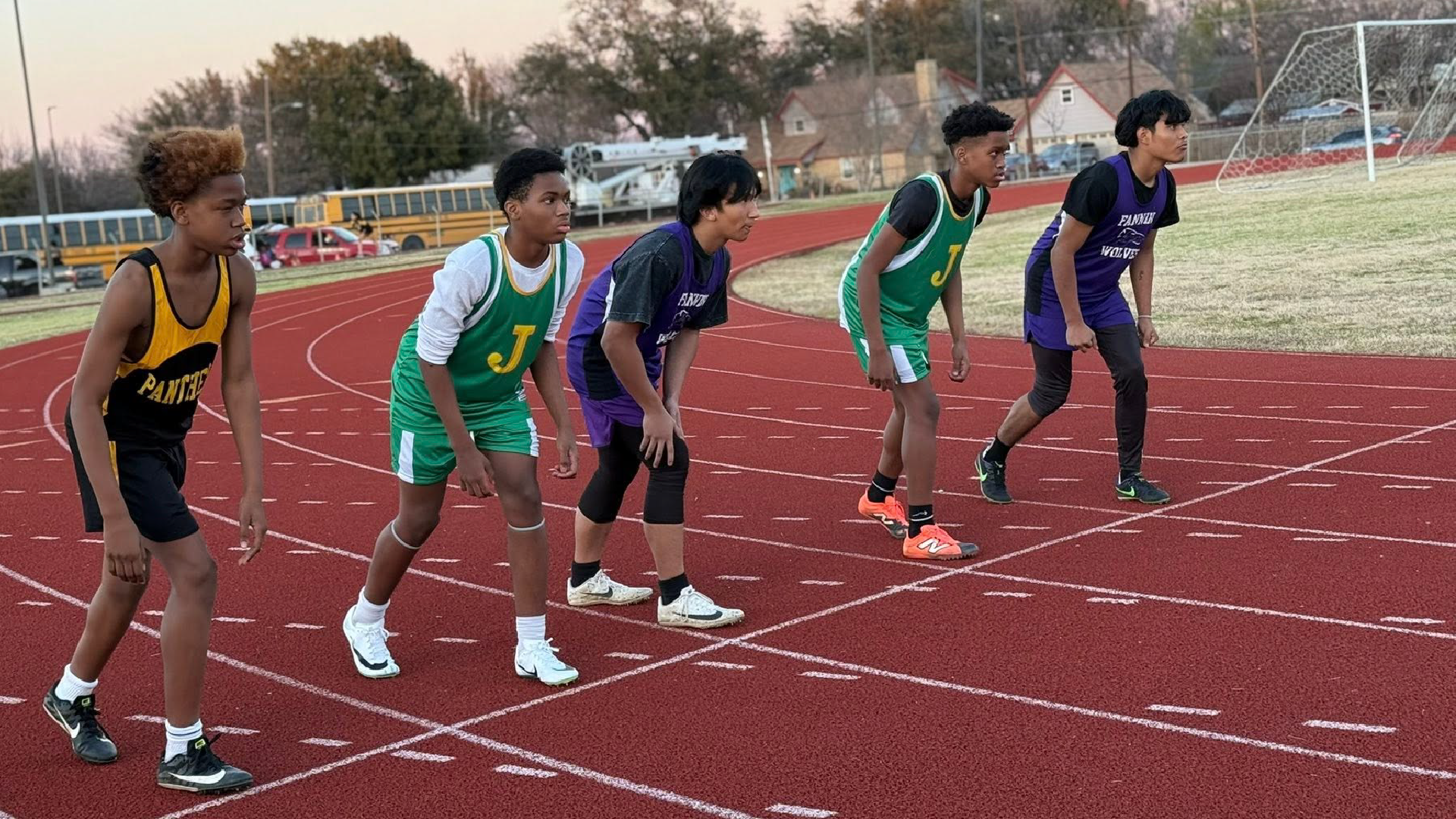 A group of young athletes in colorful athletic gear stand on a running track, with a sports field and buildings visible in the background.