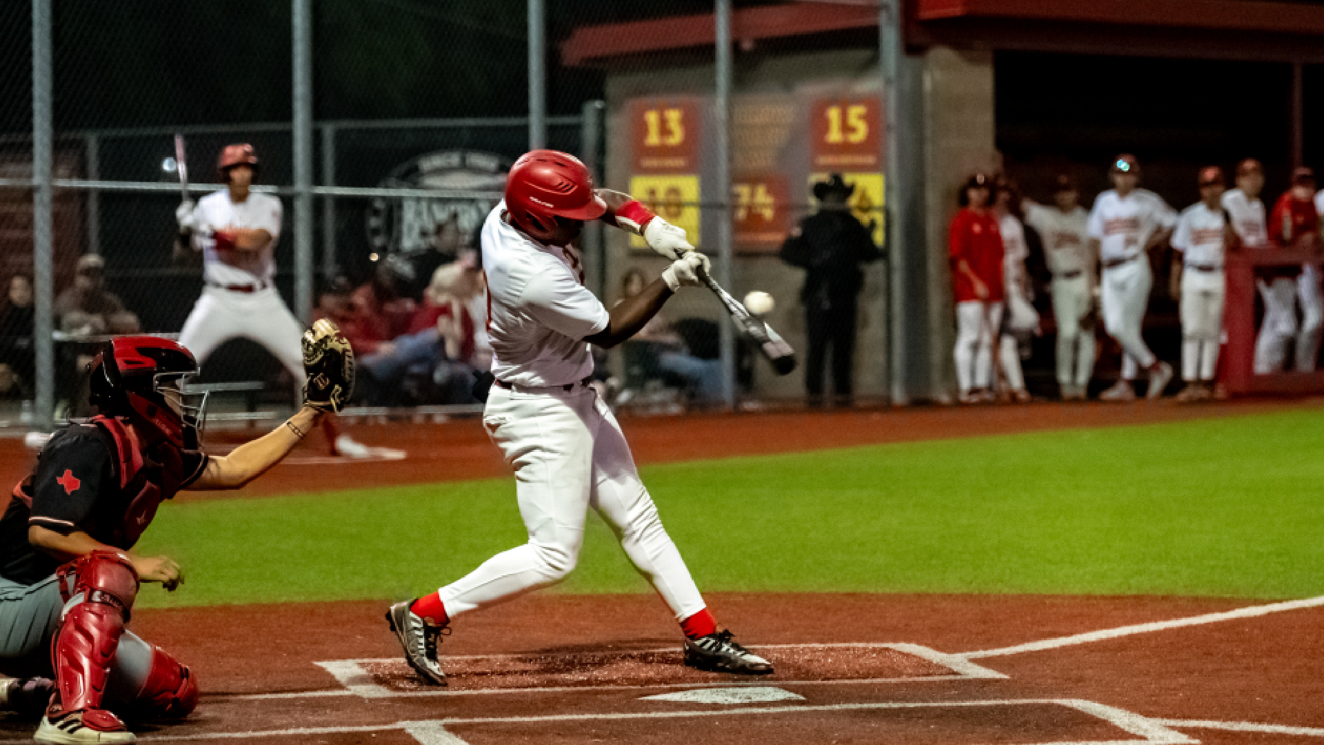 A baseball player in a white uniform stands at the plate, preparing to swing, while a catcher in a red uniform crouches behind home plate, and other players and spectators can be seen in the background.
